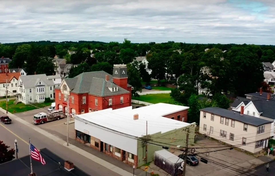 Aerial view of a small town with colorful houses, green trees, and a cloudy sky.