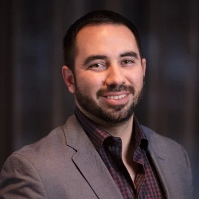 A man with dark hair and a beard, wearing a gray blazer and a dark plaid shirt, is smiling at the camera with a dark background.