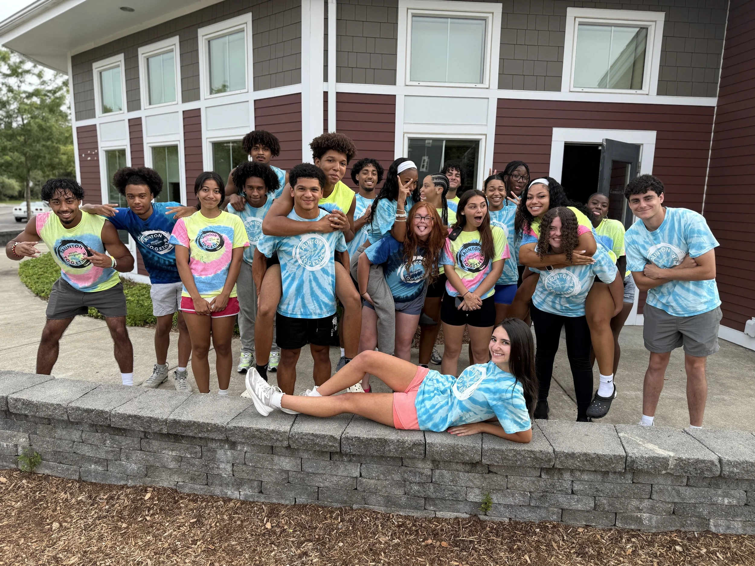 Group of teenagers wearing tie-dye t-shirts, some standing, one lying on the sidewalk, in front of a red and white building, smiling and posing for the camera.