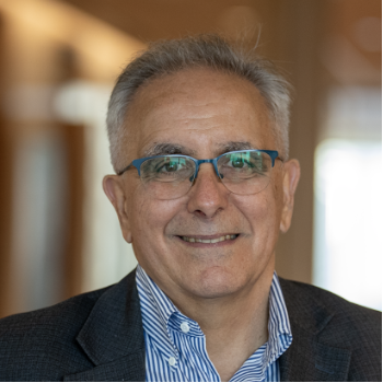 A smiling man with gray hair and glasses, wearing a dark suit and blue collared shirt, standing indoors with blurred background.