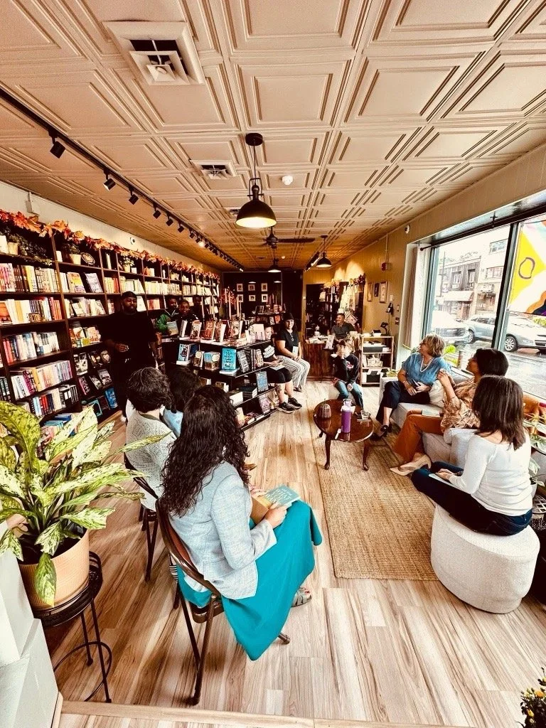 Group of people attending a book reading or discussion event inside a cozy bookstore or café with wooden floors, bookshelves, and large window letting in natural light.