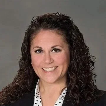 A woman with curly brown hair, smiling, wearing a black blazer and a white polka dot blouse, against a gray background.