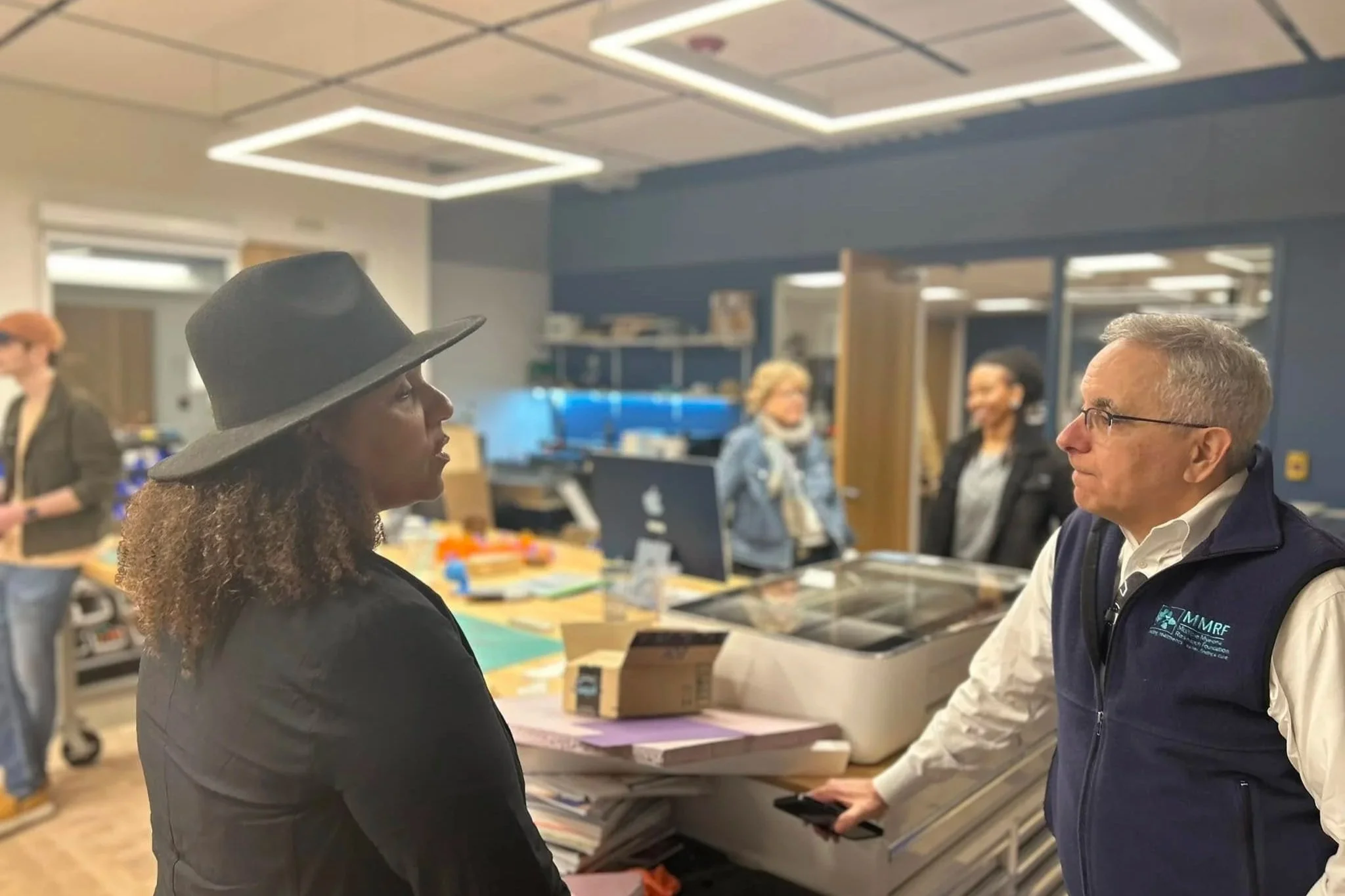 A woman wearing a black wide-brimmed hat stands in profile facing a man wearing glasses and a navy vest in an office or classroom setting, with several shelves, desks, and other people in the background.