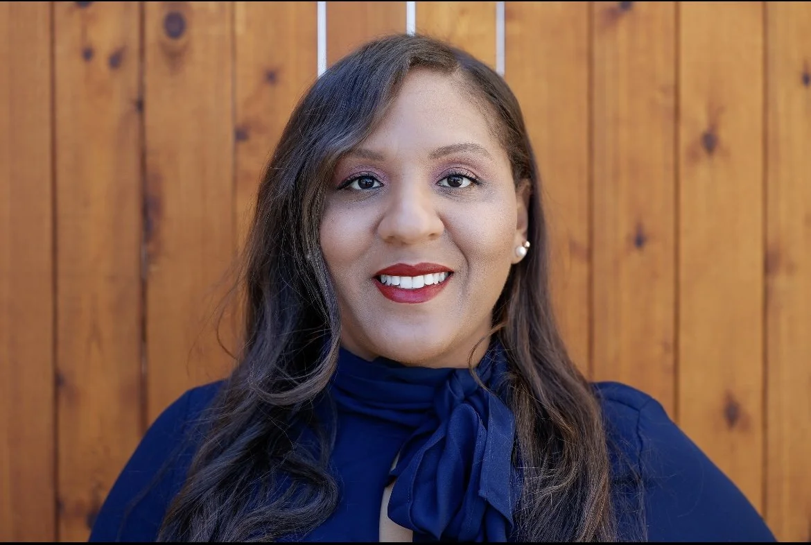 A woman with long dark hair wearing a blue top with a bow-tie detail poses in front of a wooden fence.