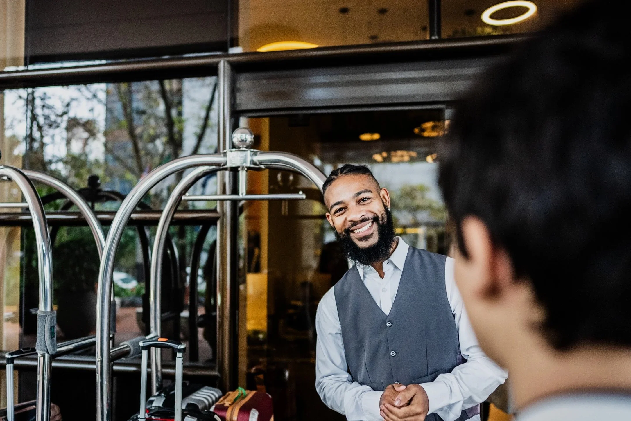 A smiling man in a vest and white shirt greeting a guest at a hotel reception desk.