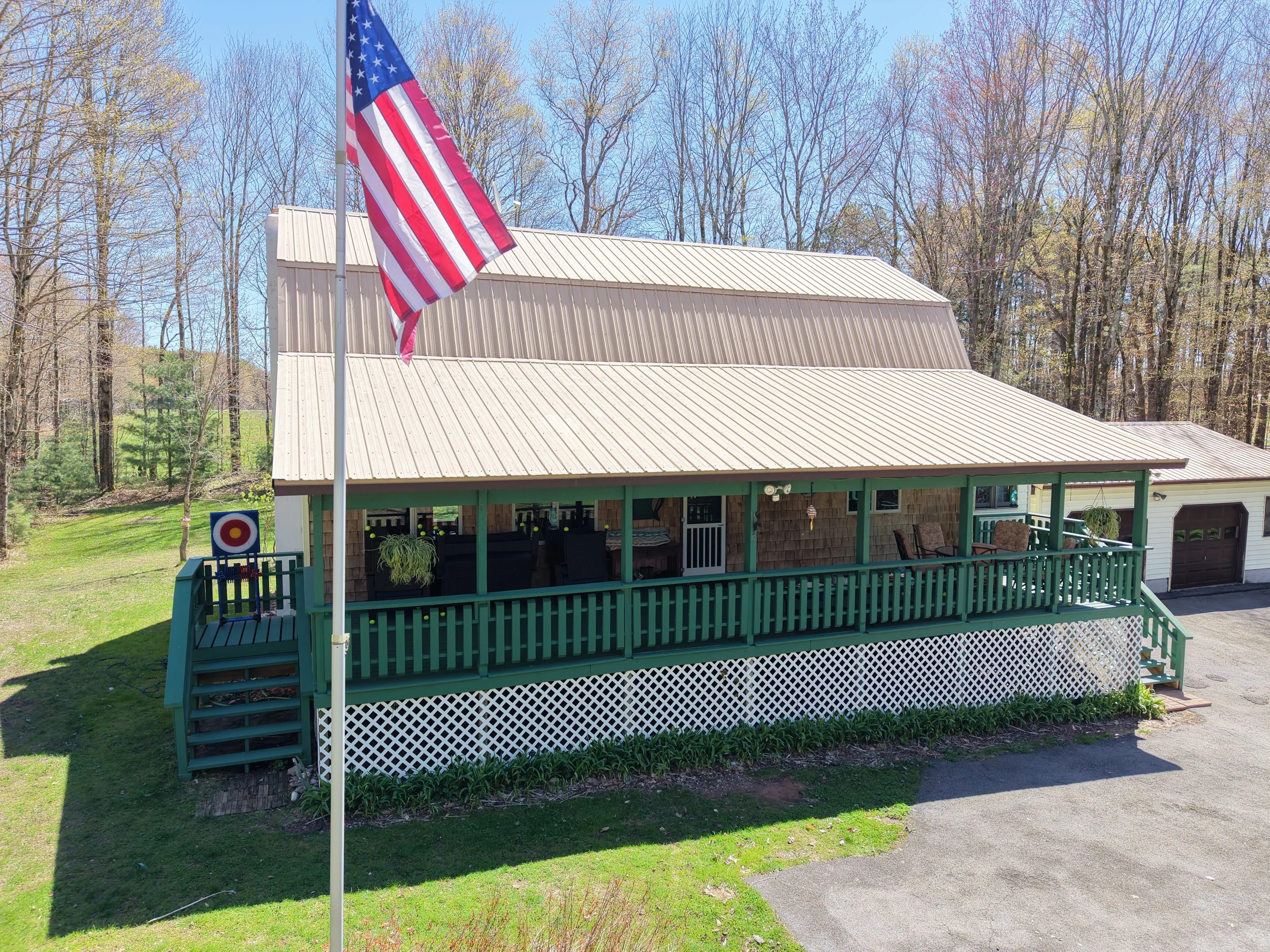 A flagpole with an American flag in front of a house with a metal roof, surrounded by trees and a green lawn, on a bright, clear day.