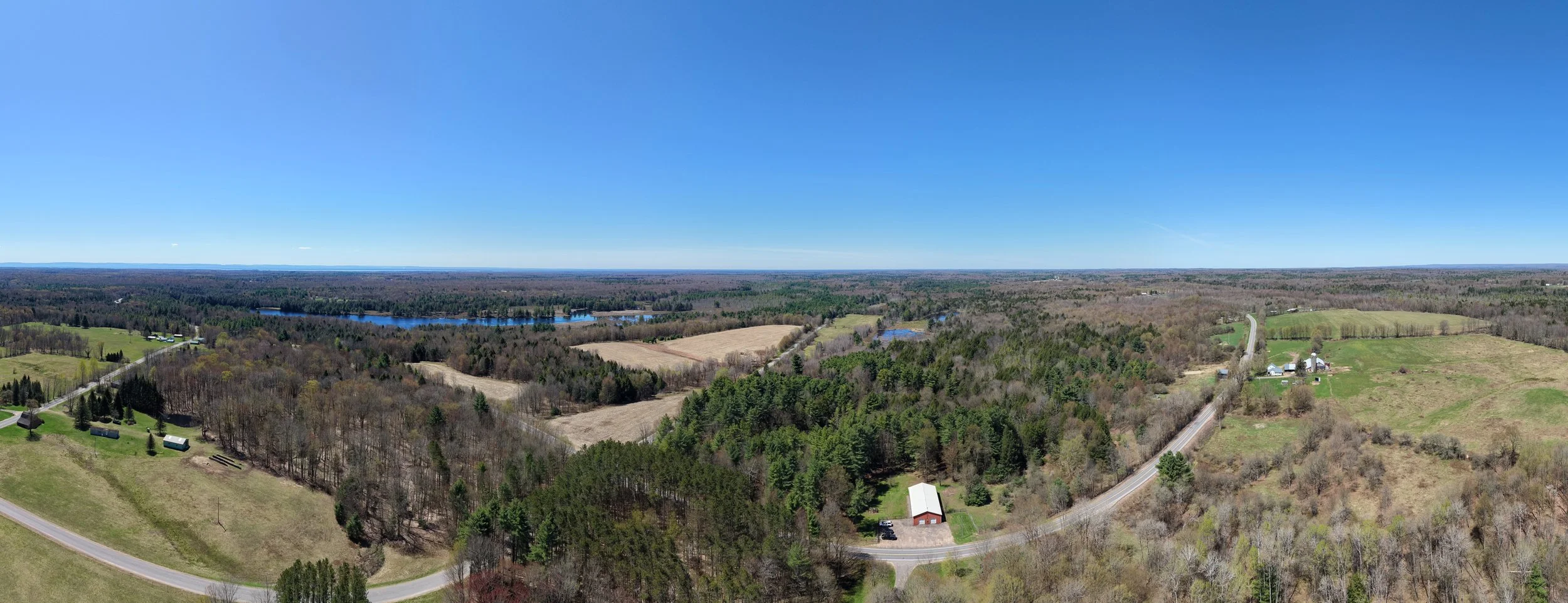 Aerial view of a rural landscape with fields, forests, lakes, and scattered farmhouses under a clear blue sky.