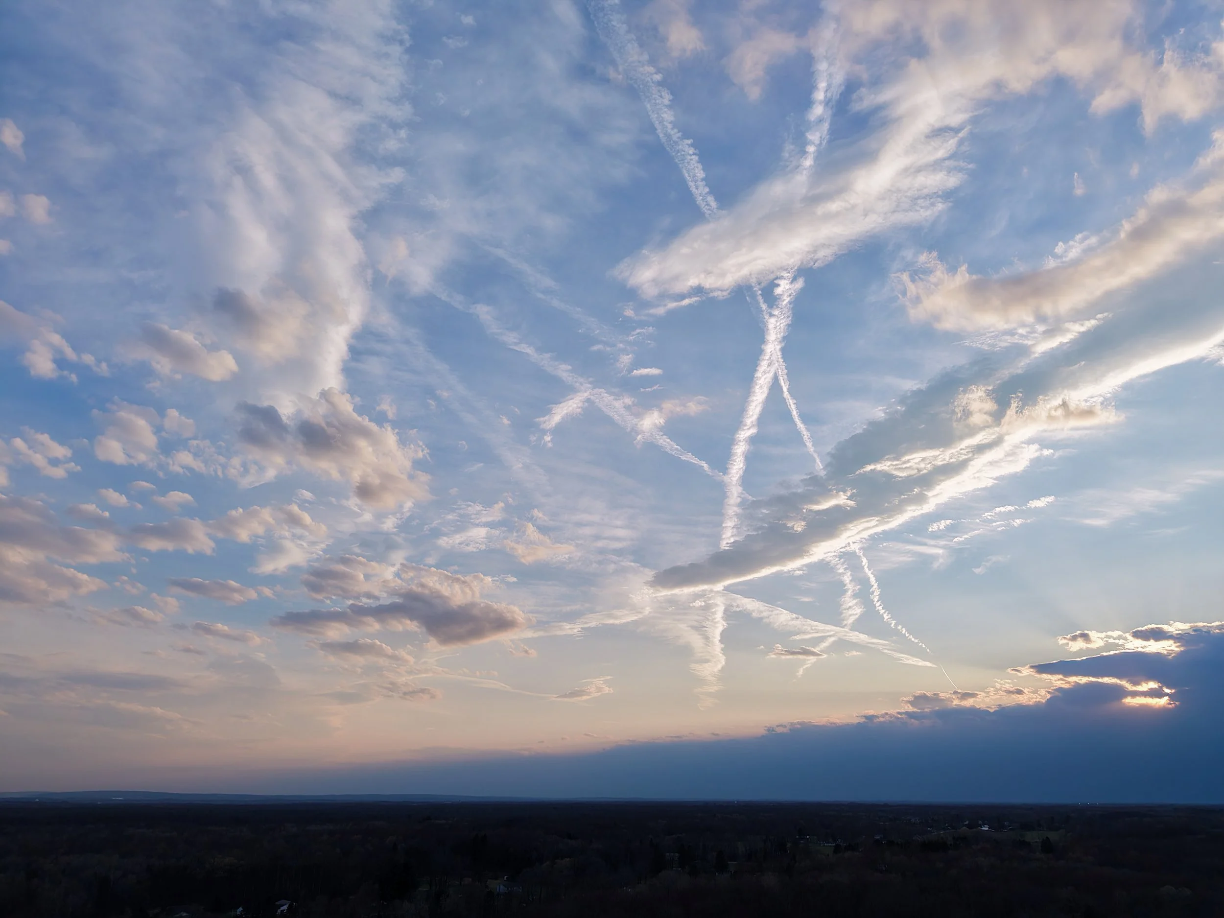 Sky with scattered clouds and multiple aircraft contrails crossing, during sunset or sunrise over a landscape.