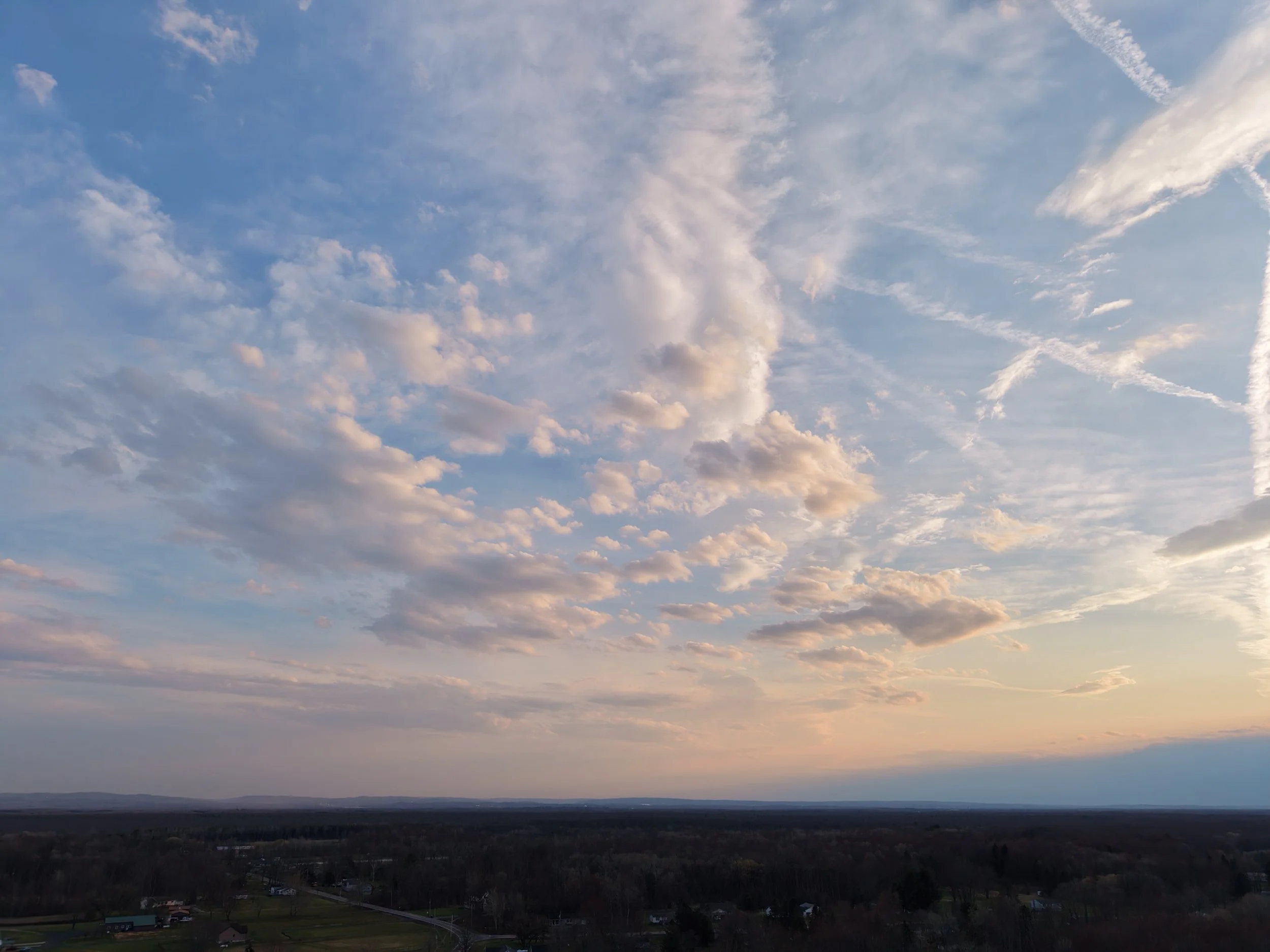 A wide view of a sky with scattered clouds during sunset over a flat landscape with trees and a few buildings.