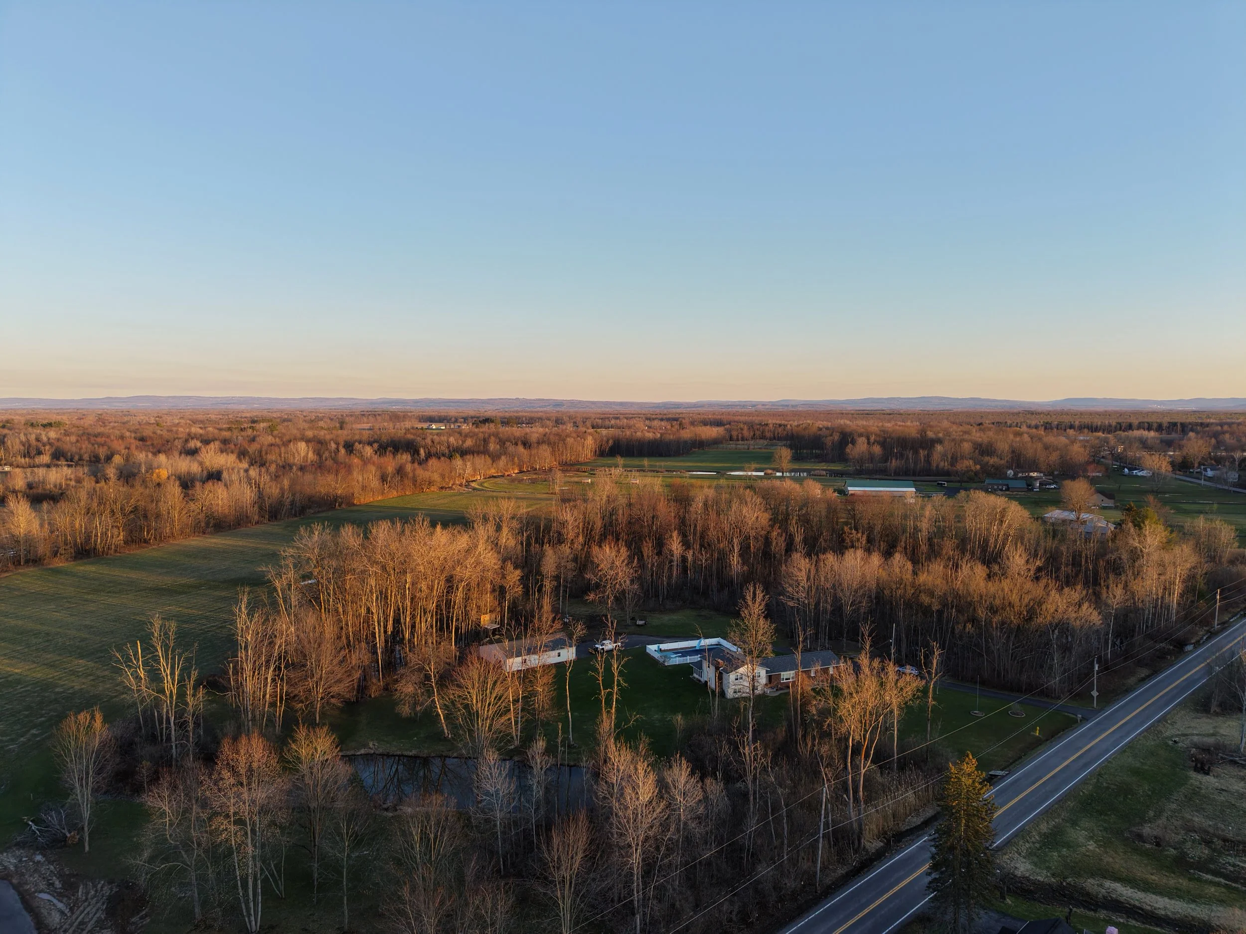 Aerial view of a rural landscape with fields, trees, a pond, houses, and a road under a clear sky at sunset.