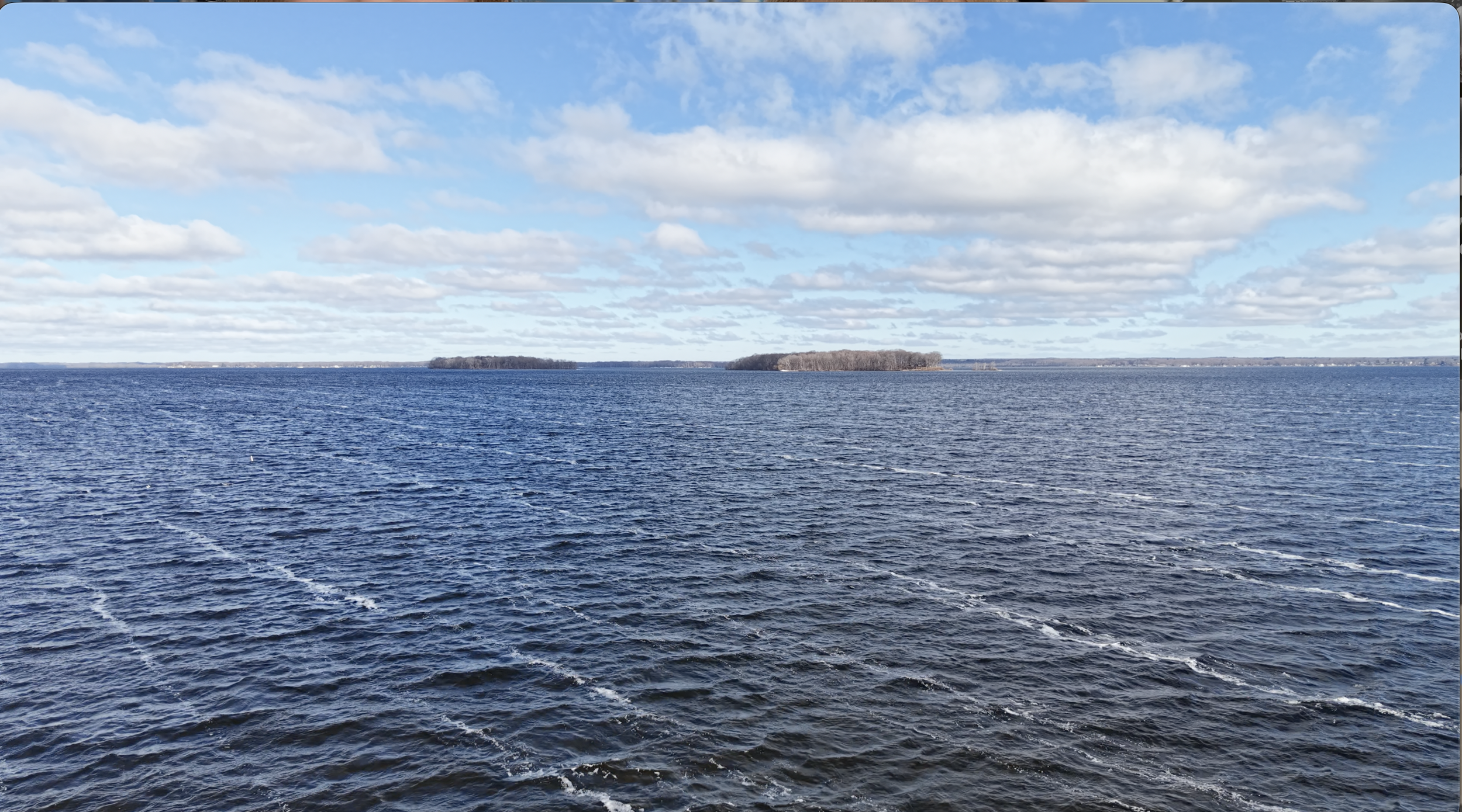 A large body of water with gentle waves, two small islands in the distance, and a partly cloudy sky overhead.