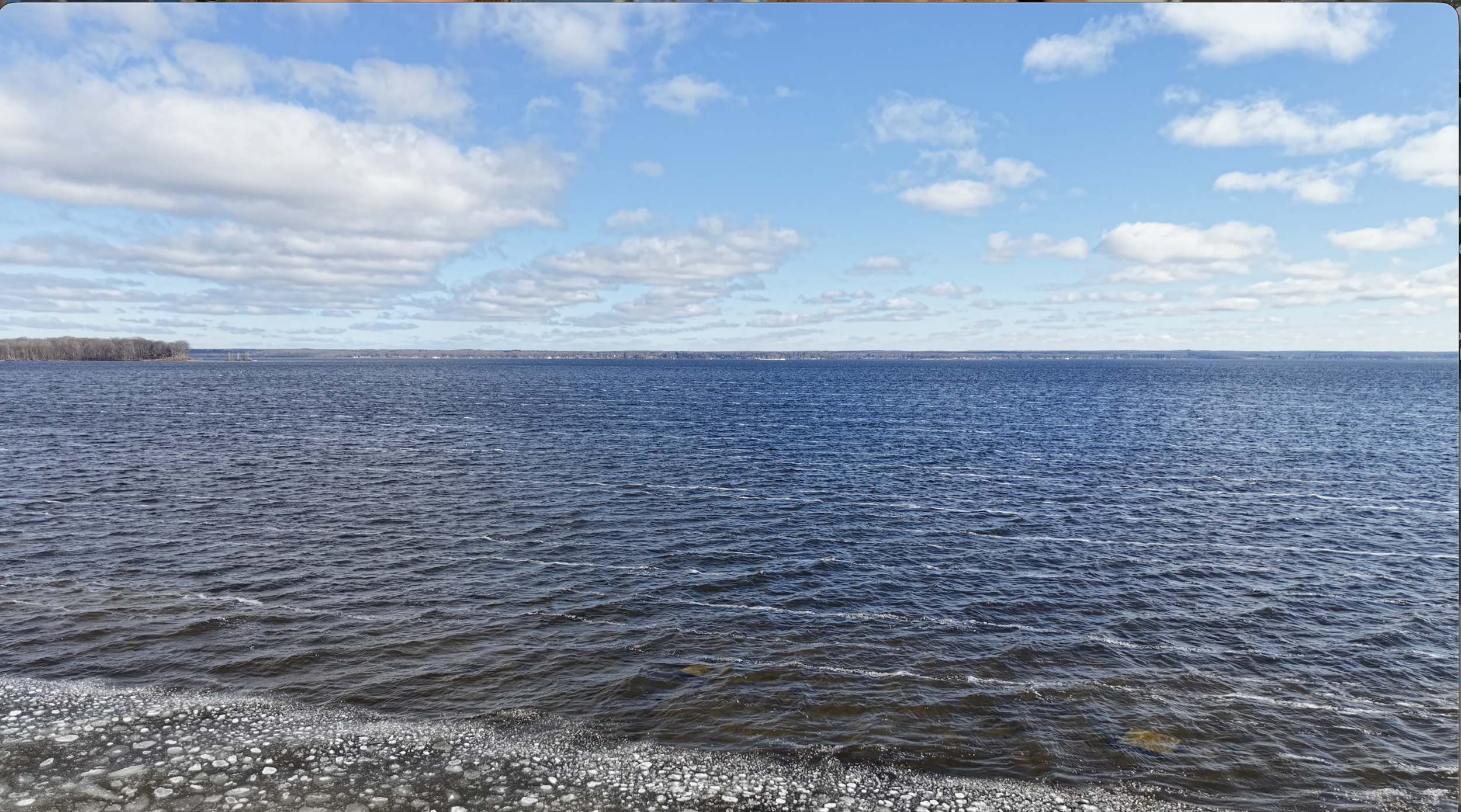 A large body of water, possibly a lake or ocean, with trees in the distance under a partly cloudy sky.