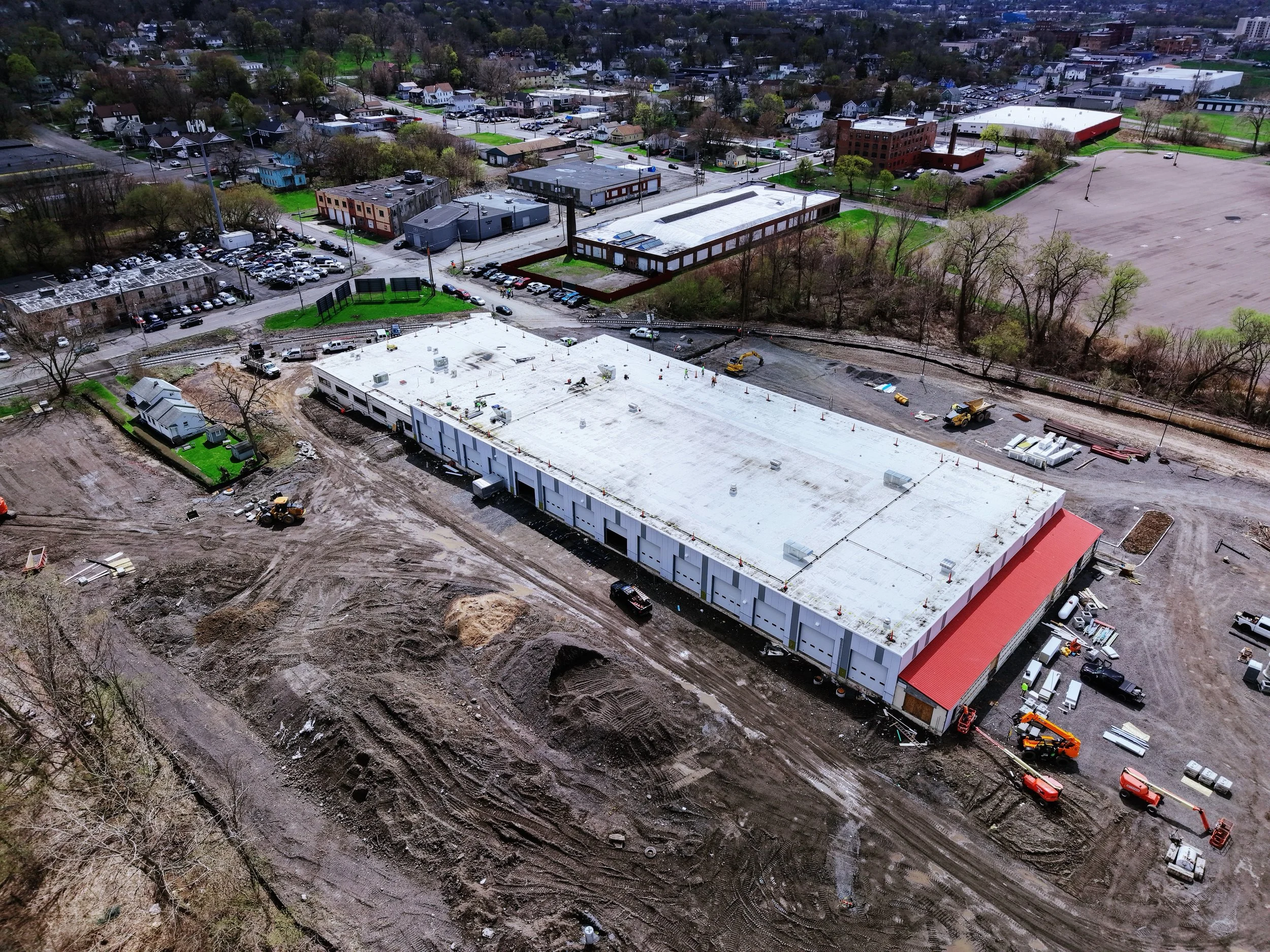 An aerial view of a construction site with large building foundations being developed, surrounded by parked cars, roads, trees, and nearby buildings in a city neighborhood.