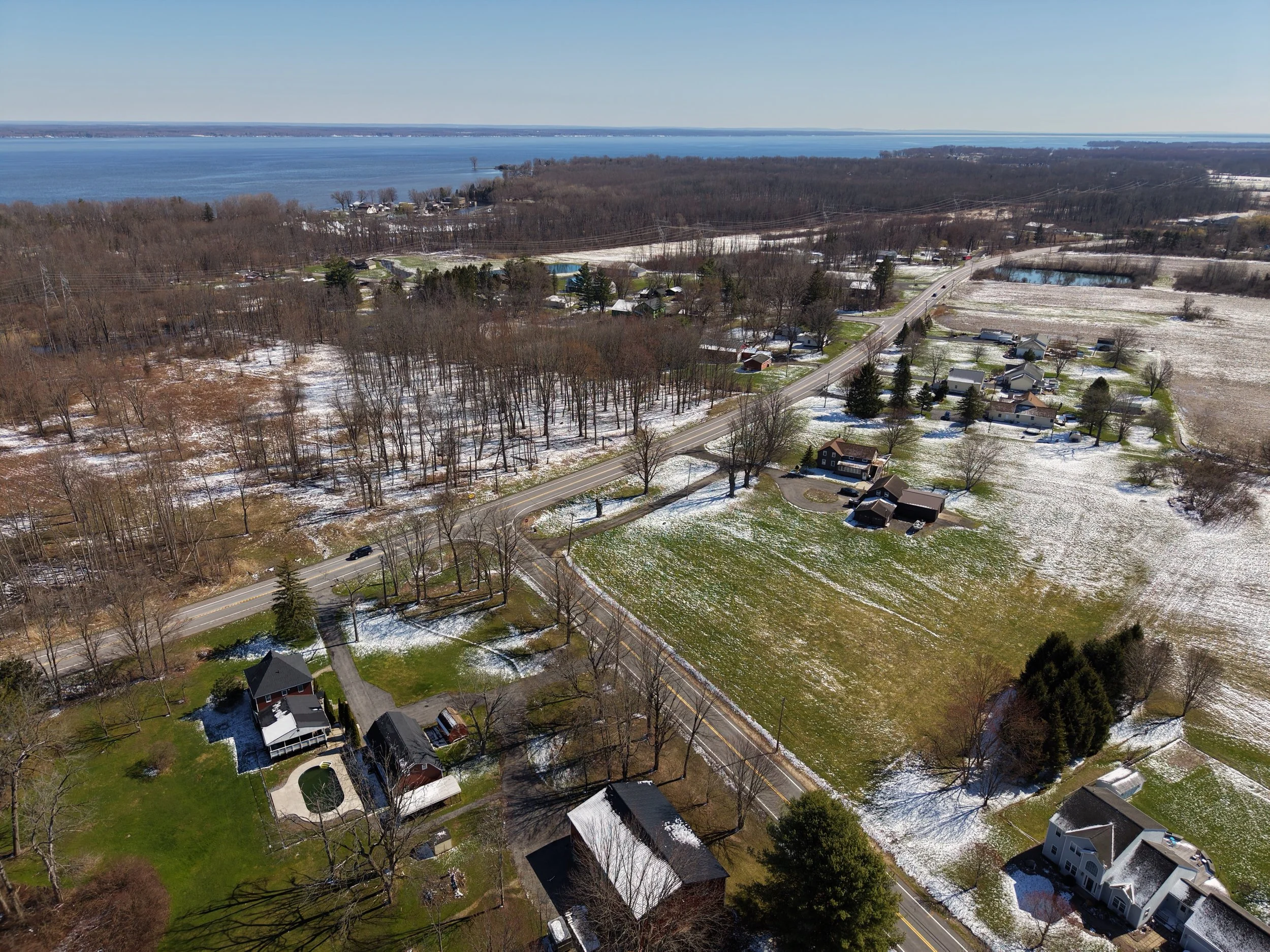 An aerial view of a suburban neighborhood with houses, trees, and roads, some patches of snow on the ground, near a large lake in the background on a clear day.