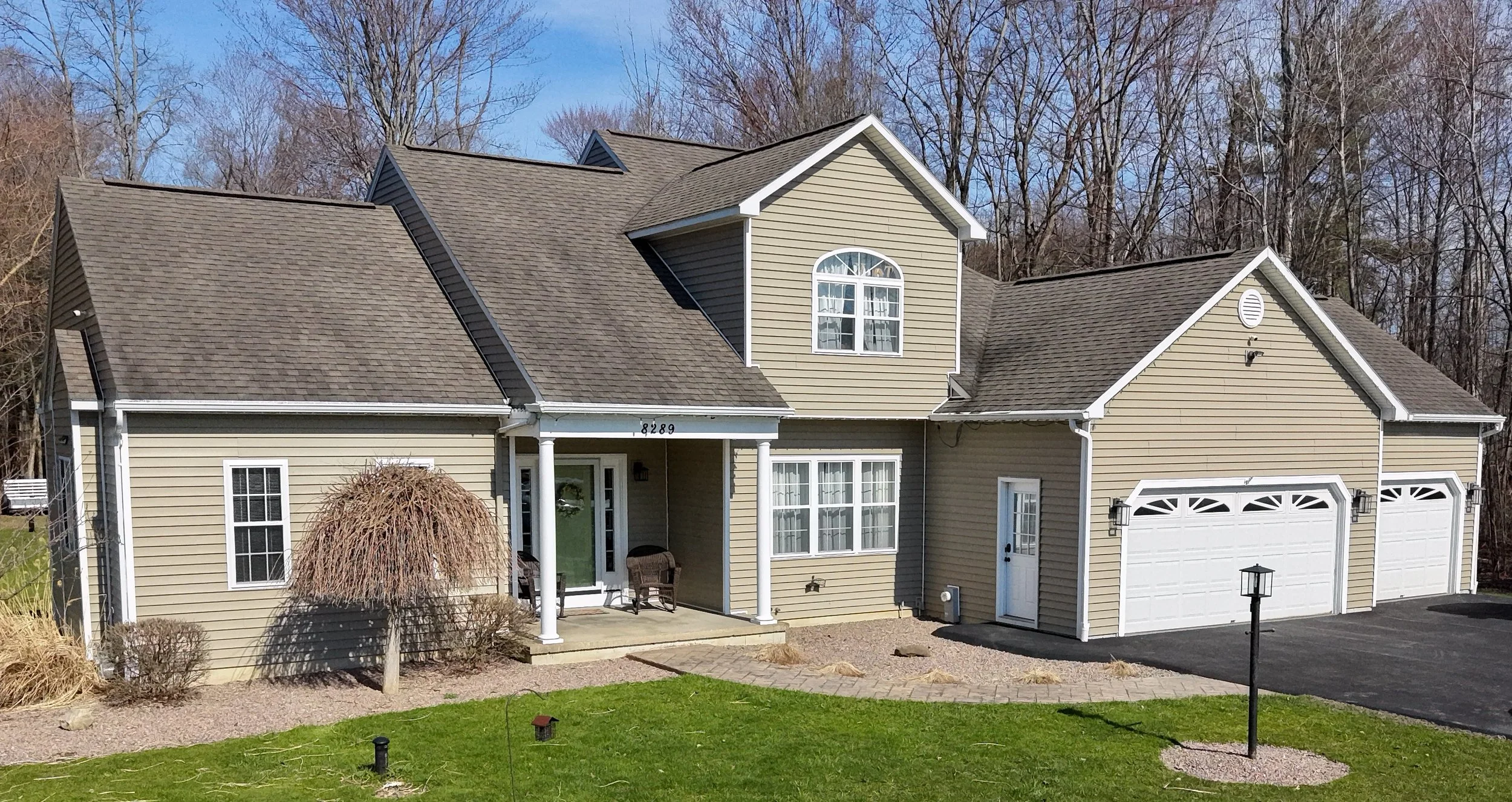 A two-story beige house with a three-car garage, a small front porch, and a manicured lawn, with leafless trees in the background.