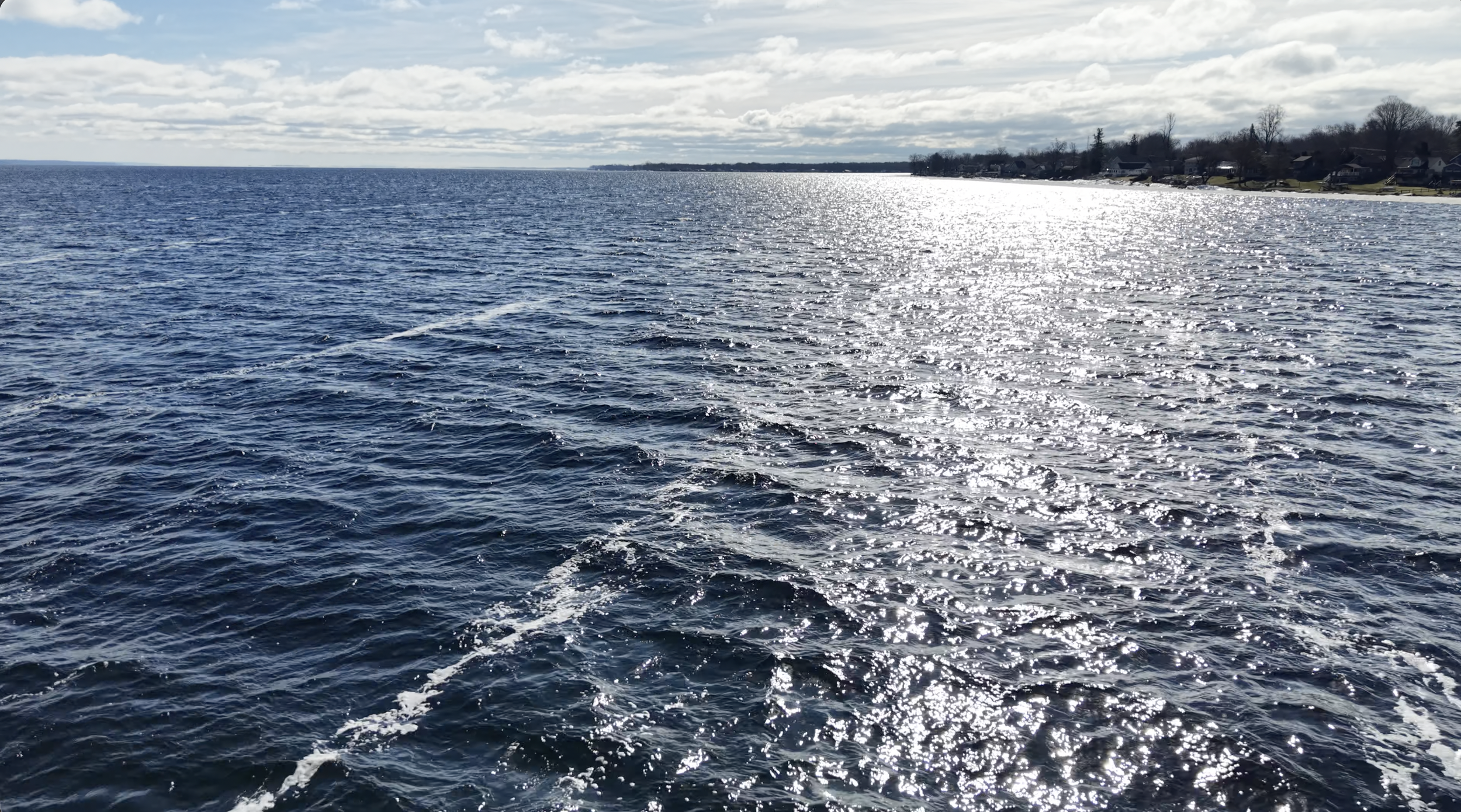 A large body of water reflecting sunlight with houses and trees along the distant shoreline under a partly cloudy sky.
