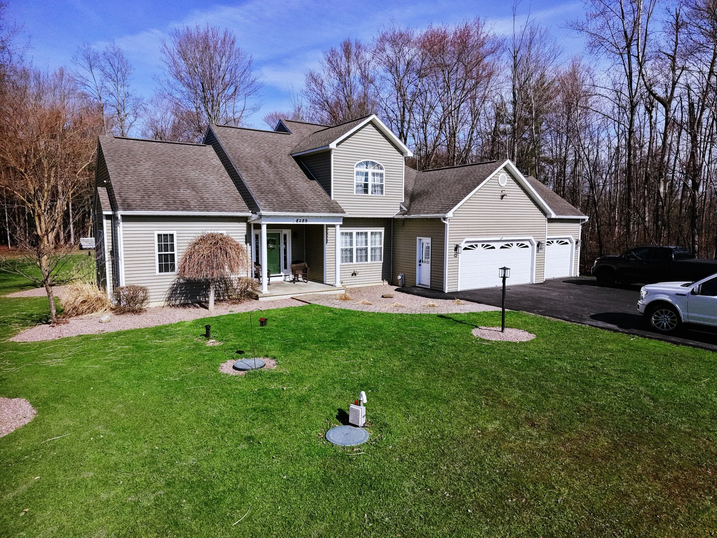A two-story house with beige siding, white trim, and a dark shingle roof. It has a front porch with seating, a garage, and a driveway with parked cars. There is a well-maintained front lawn, small trees, and a wooded background.