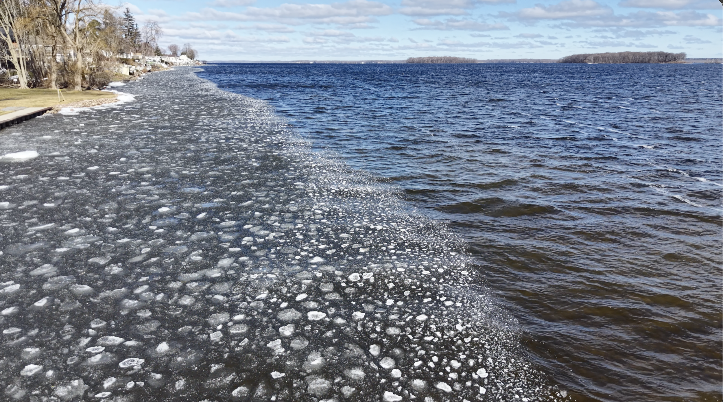 Frozen shoreline with ice cover near houses and trees, water on the right, cloudy sky in the background.