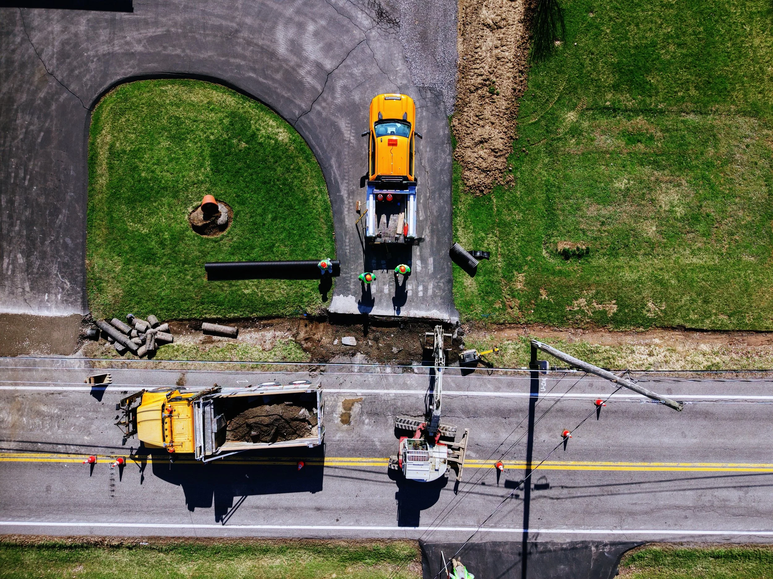 An aerial view of road construction work with several workers, trucks, and construction equipment on a street next to grassy areas.