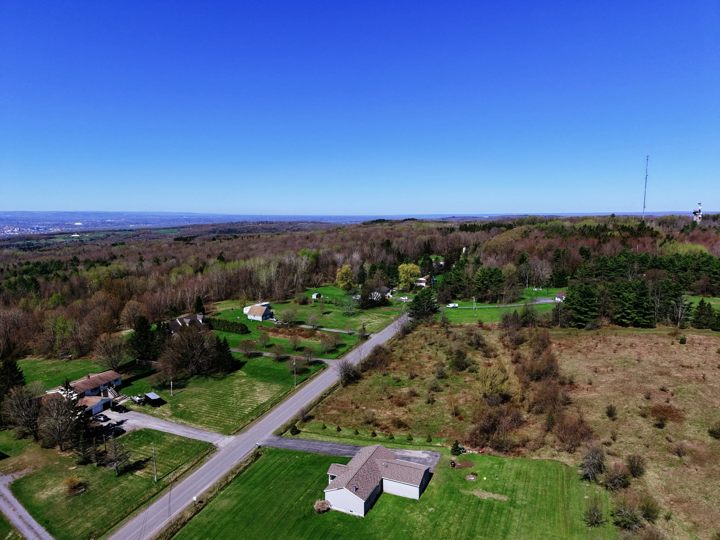 Aerial view of a suburban landscape with houses, trees, and a large wooded area under a clear blue sky.