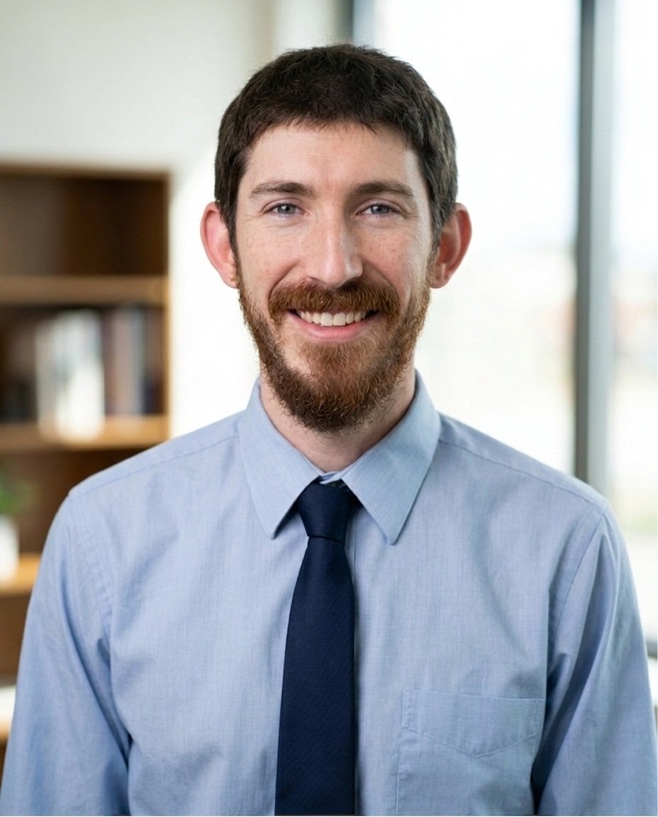 A smiling man with brown hair, beard, and mustache wearing a light blue dress shirt and dark blue tie, standing indoors near a window.