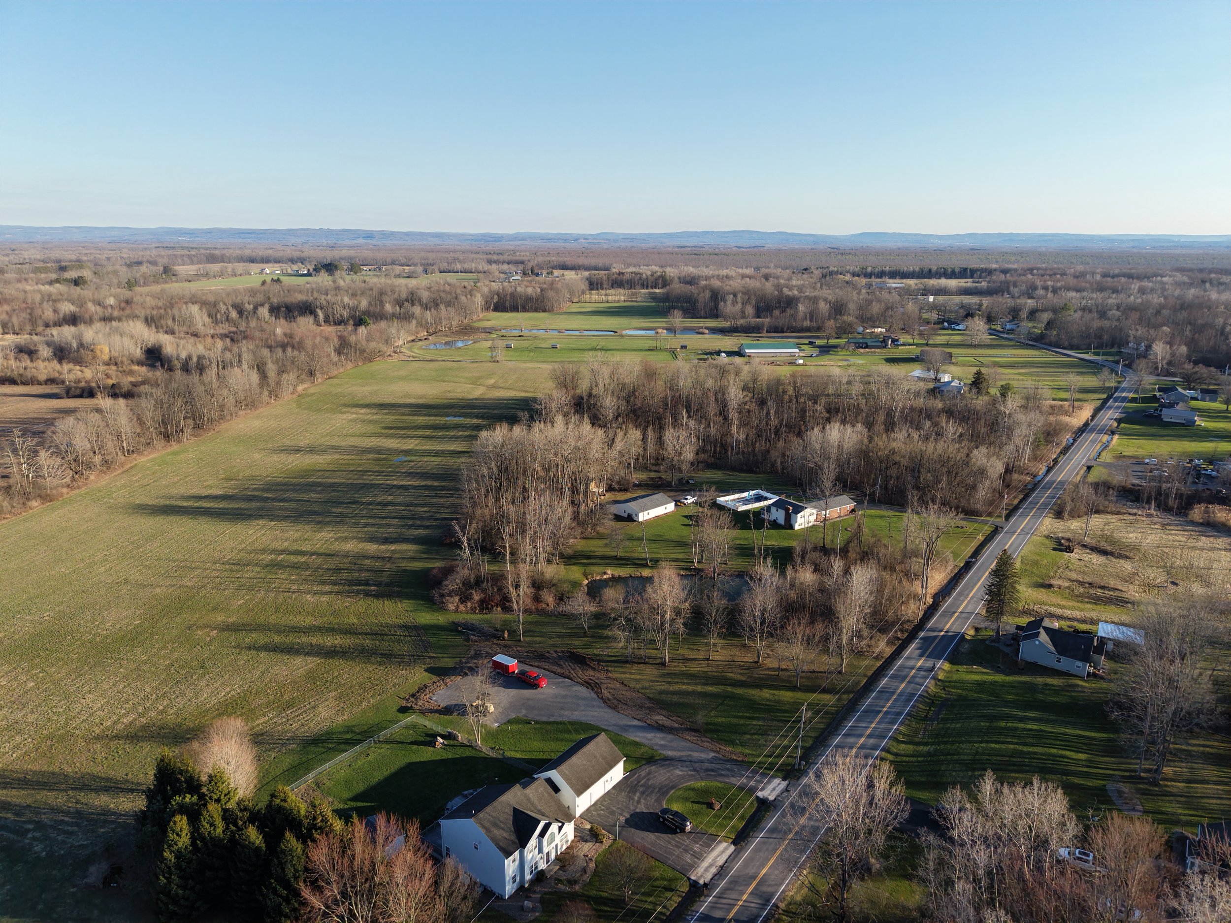 A rural landscape with open fields, houses, trees, and a road stretching into the distance under a clear sky.