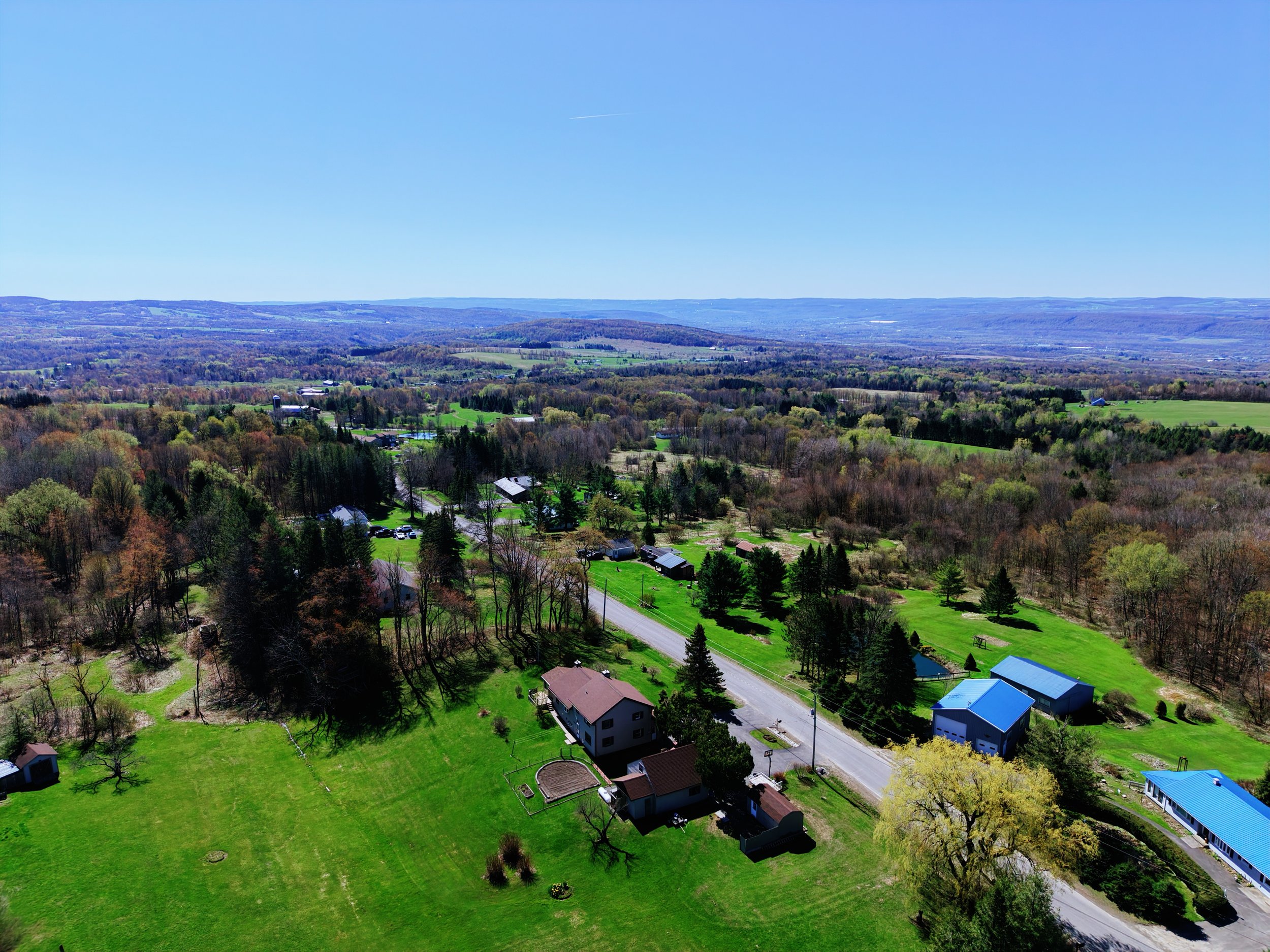 A scenic view of a rural landscape with rolling hills, numerous trees, scattered houses, and open green fields under a clear blue sky.