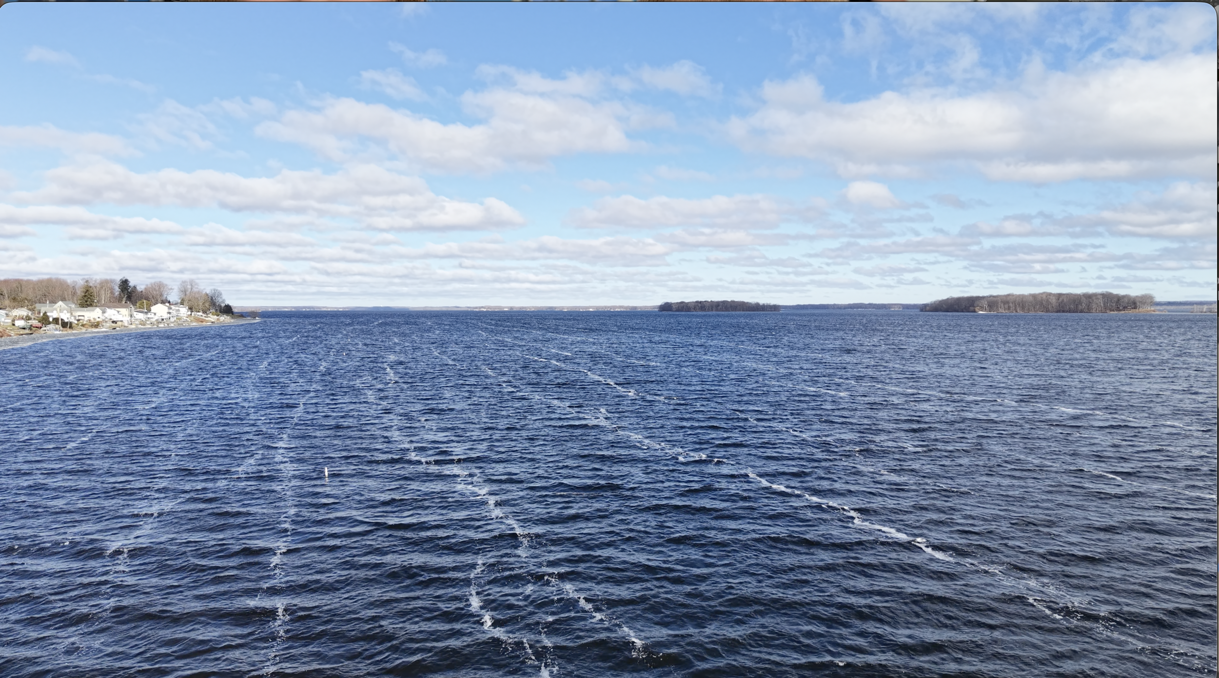 View of a large body of water with a wake trail from a boat, houses on the shoreline to the left, and small distant islands on the horizon under a partly cloudy sky.