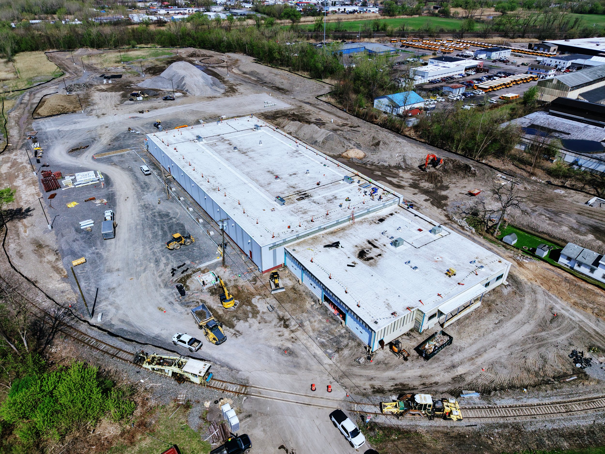 Aerial view of a construction site with a flat white roof, construction vehicles, and materials, surrounded by roads, trees, and nearby buildings, including a parking lot with yellow school buses.