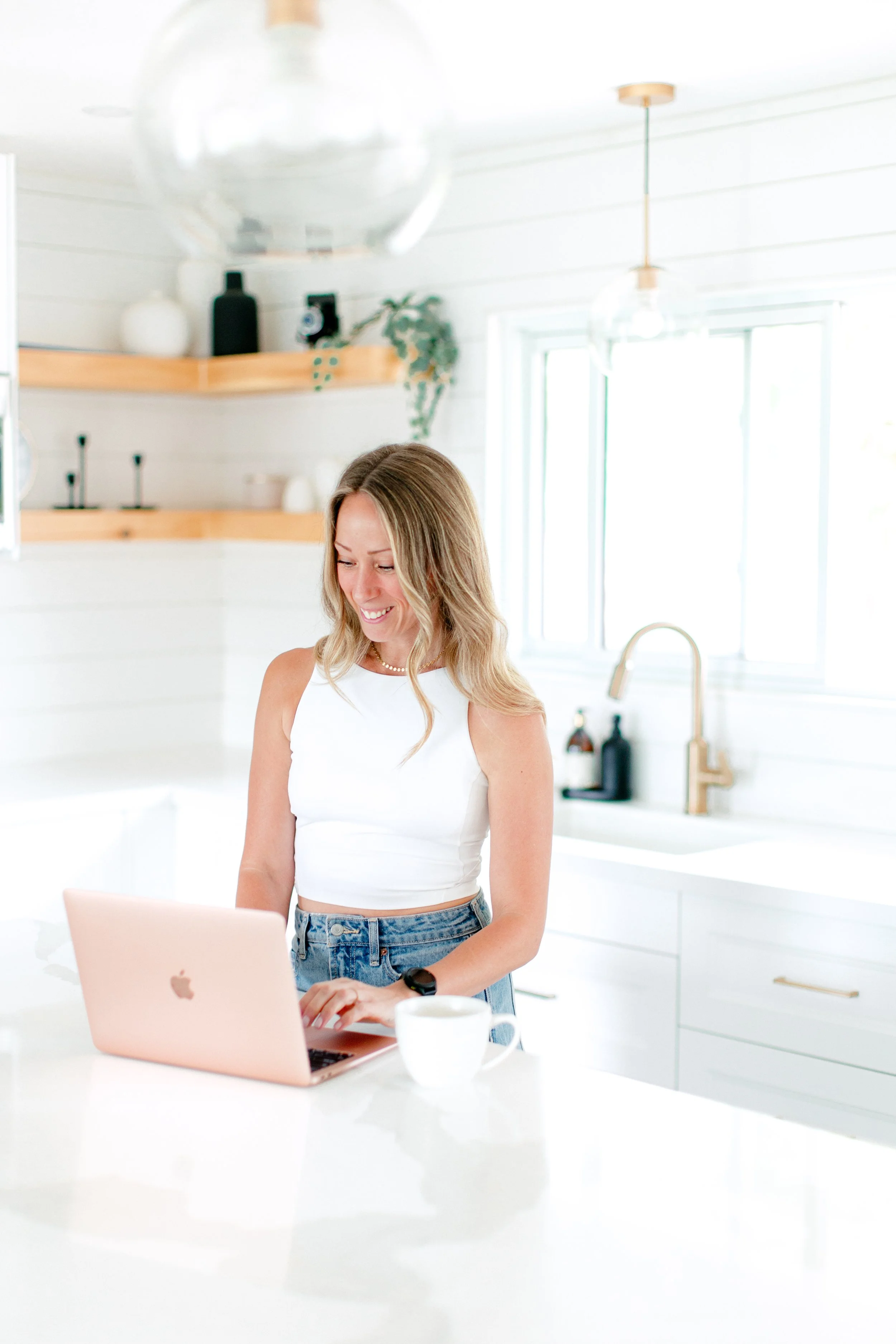 A woman smiling while using a pink laptop in a bright, modern kitchen with white cabinets and light wood accents.
