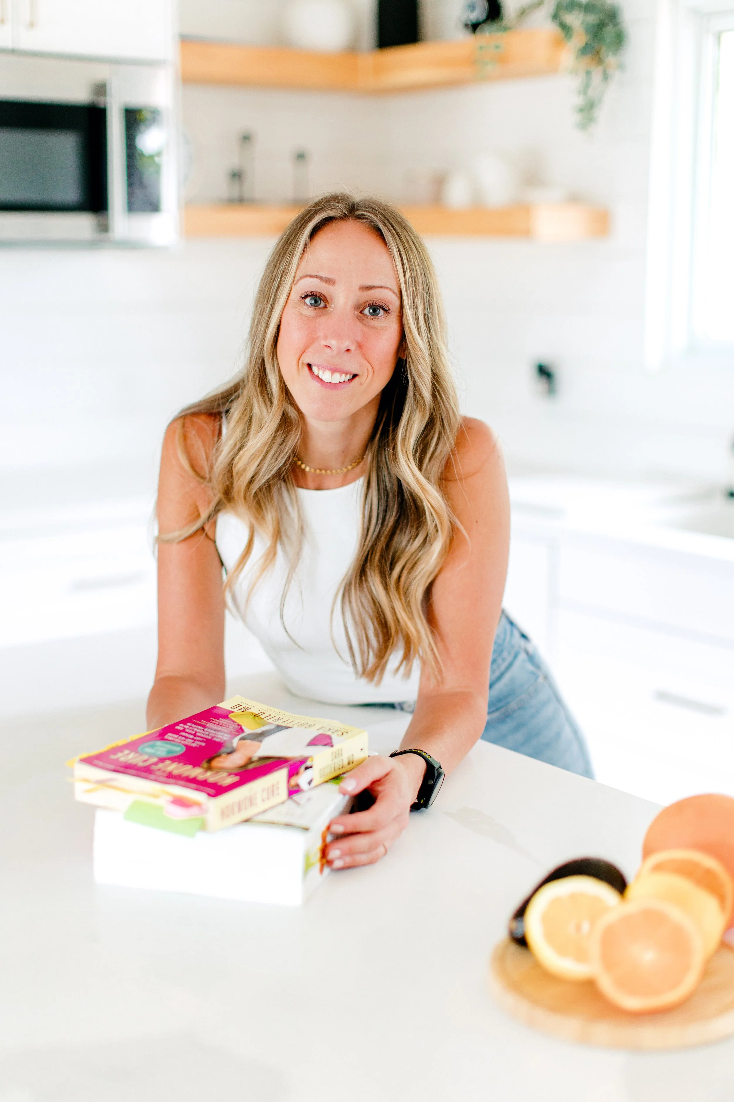 A woman with long blonde hair smiling, leaning on a kitchen counter with books in front of her and a platter of sliced lemons, oranges, and an avocado on the side.