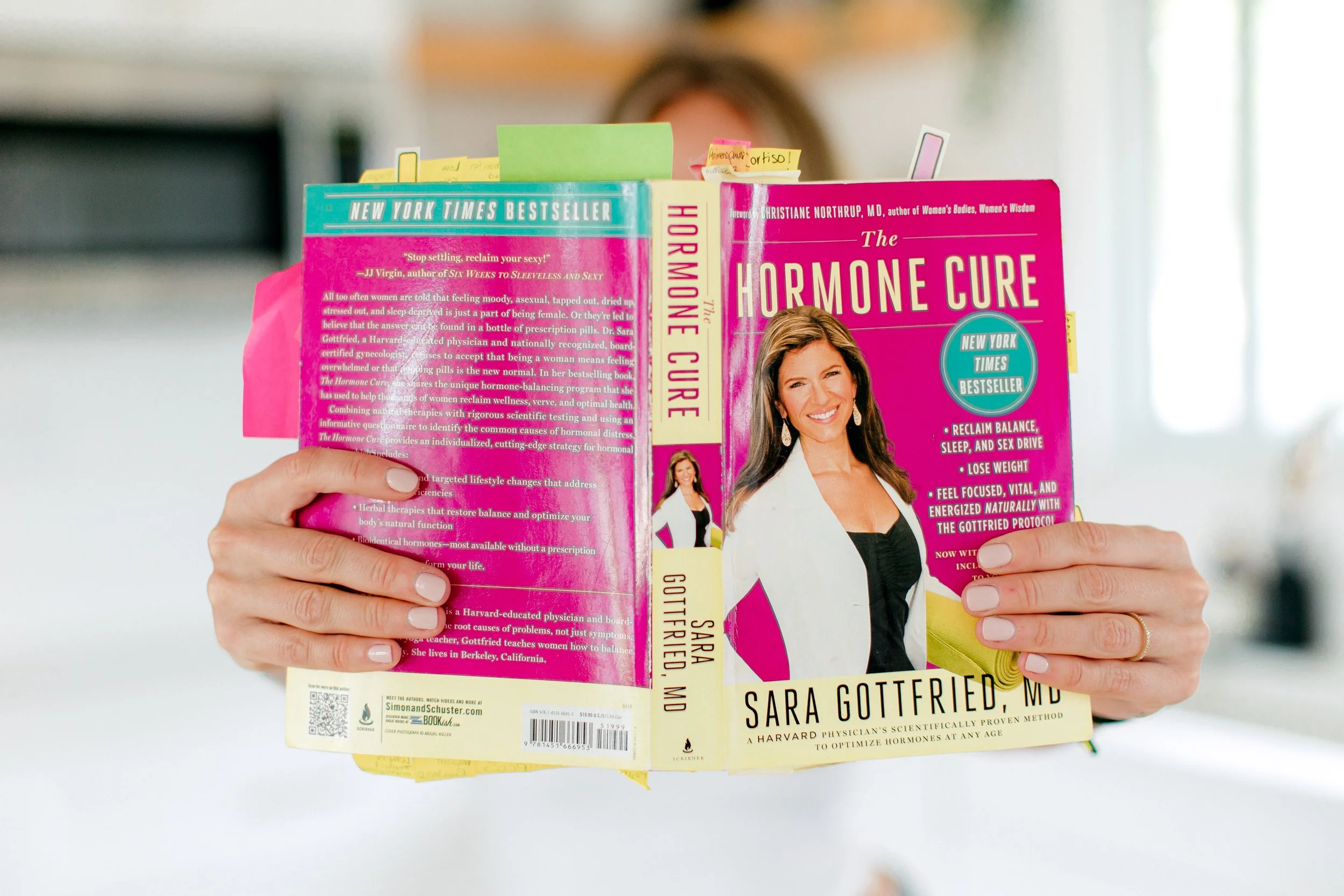 A person holding a pink-colored book titled "The Hormone Cure" by Sara Gottfried, MD, with colorful sticky notes. The book features a smiling woman on the cover and is a New York Times bestseller about balancing hormones, sleep, weight loss, and natural health solutions.