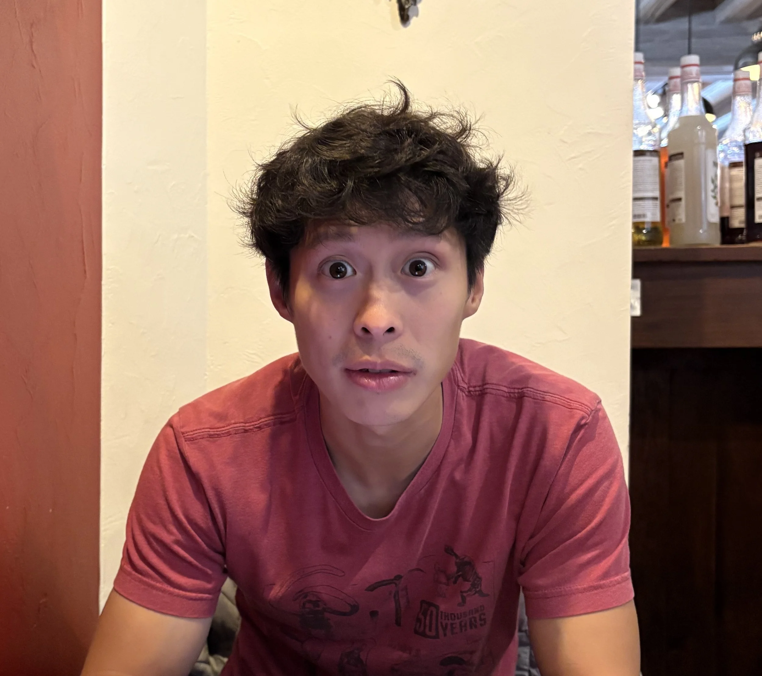 A young man with dark curly hair and wide eyes looking surprised or confused, sitting in a cozy cafe or restaurant with red and white walls, and a shelf with bottles behind him.