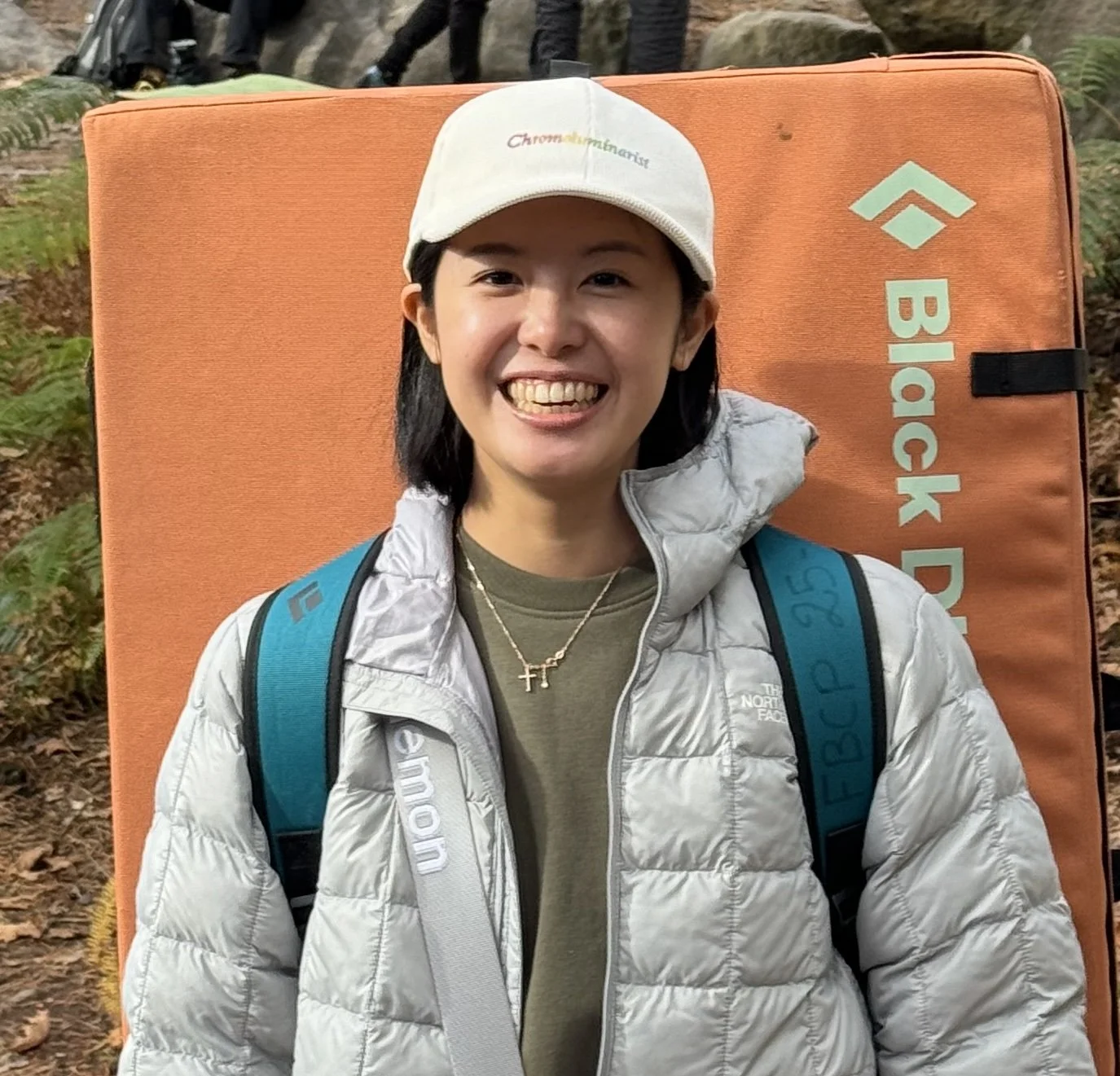 A smiling woman with short black hair wearing a white cap, gray North Face jacket, olive green shirt, and a necklace with a cross pendant, standing in front of a large orange climbing pad with the words "Black Diamond" on it, outdoors with a forest background.