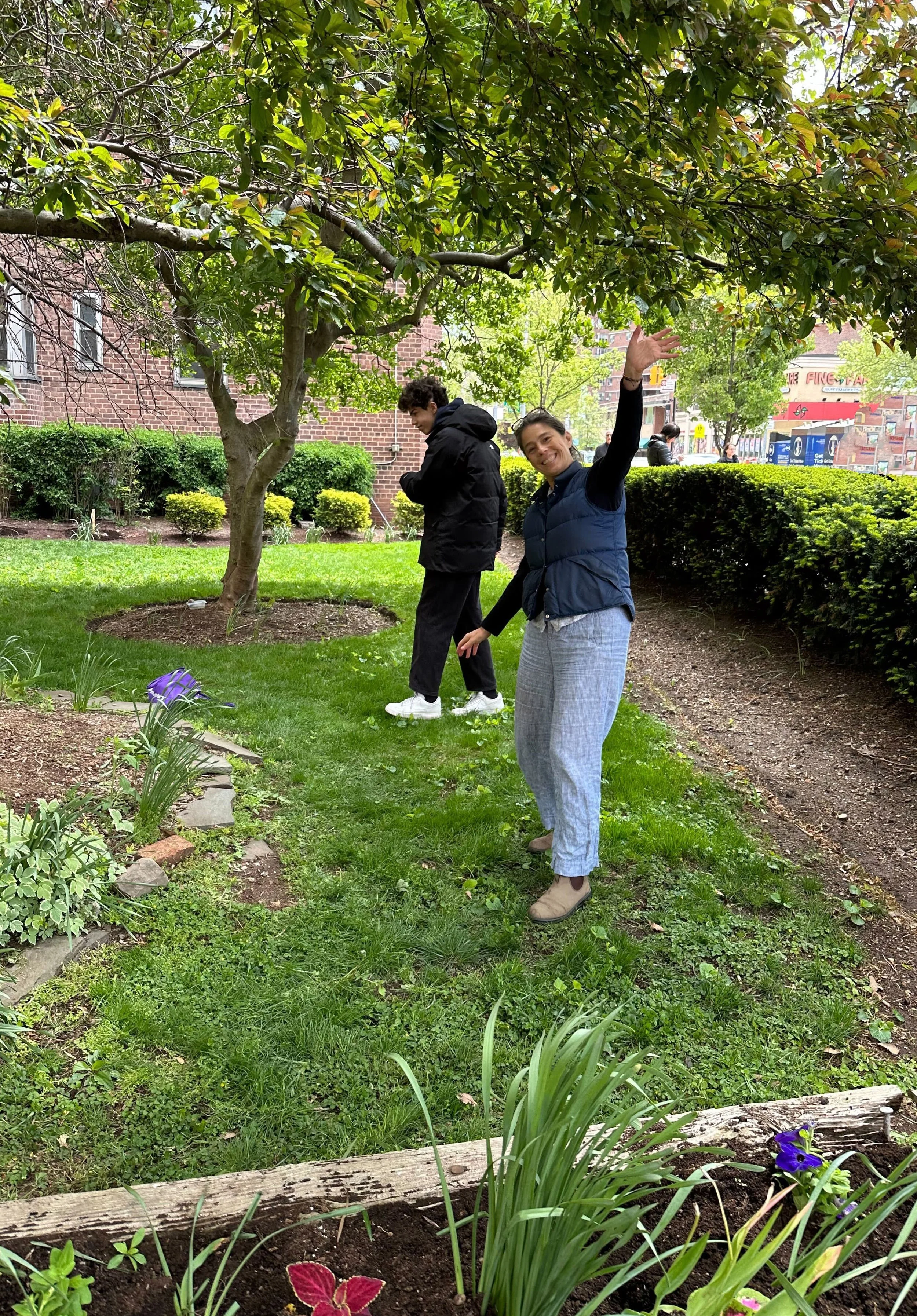 Two people smiling and gesturing under a tree outside a brick building