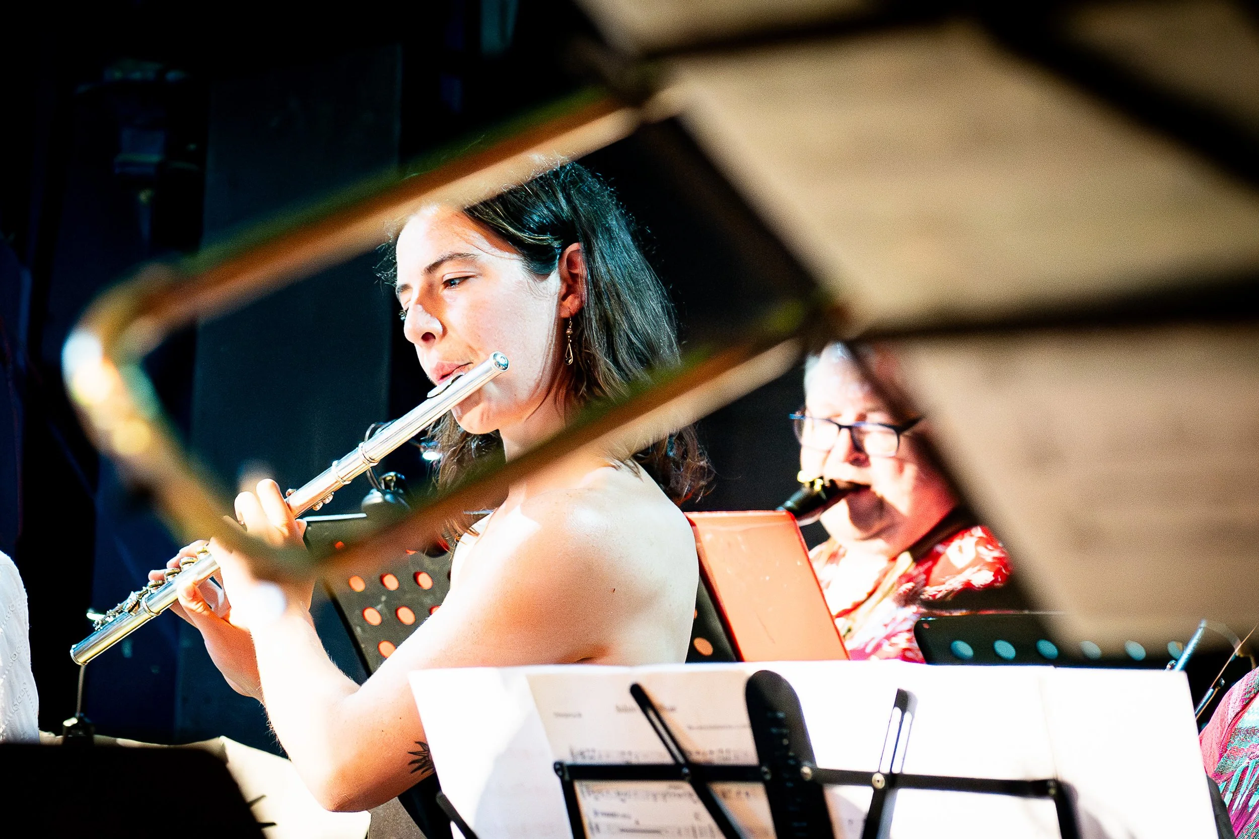 Musicians performing on stage, with a woman playing a flute in the foreground and another musician playing a wind instrument in the background.