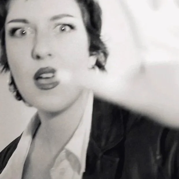 A woman with short dark hair, wearing a white shirt and dark jacket, making a surprised or confused facial expression.