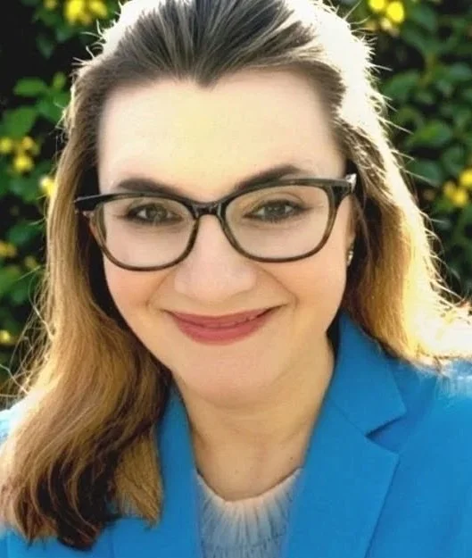 A woman with long brown hair, wearing glasses, a blue blazer, and a white sweater, smiling outdoors with greenery in the background.