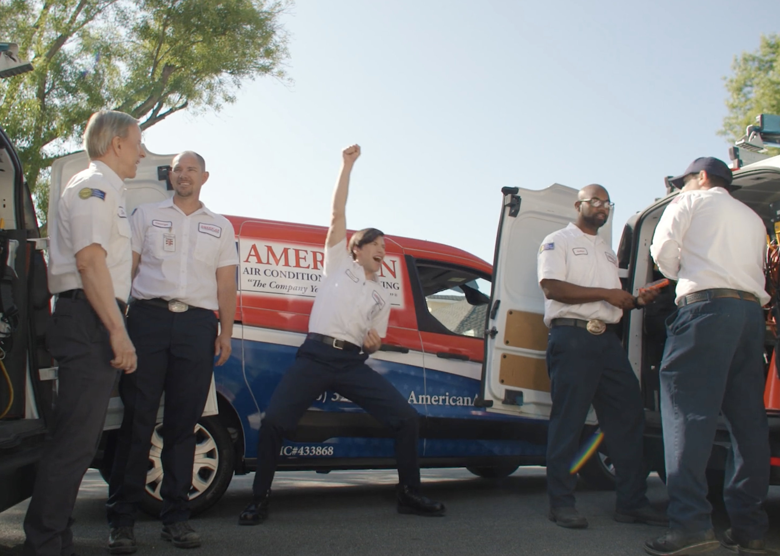 Group of people standing next to service trucks, some with open tool compartments, engaging in conversation or tasks outdoors with trees and clear sky in background, possibly during a work or training event.