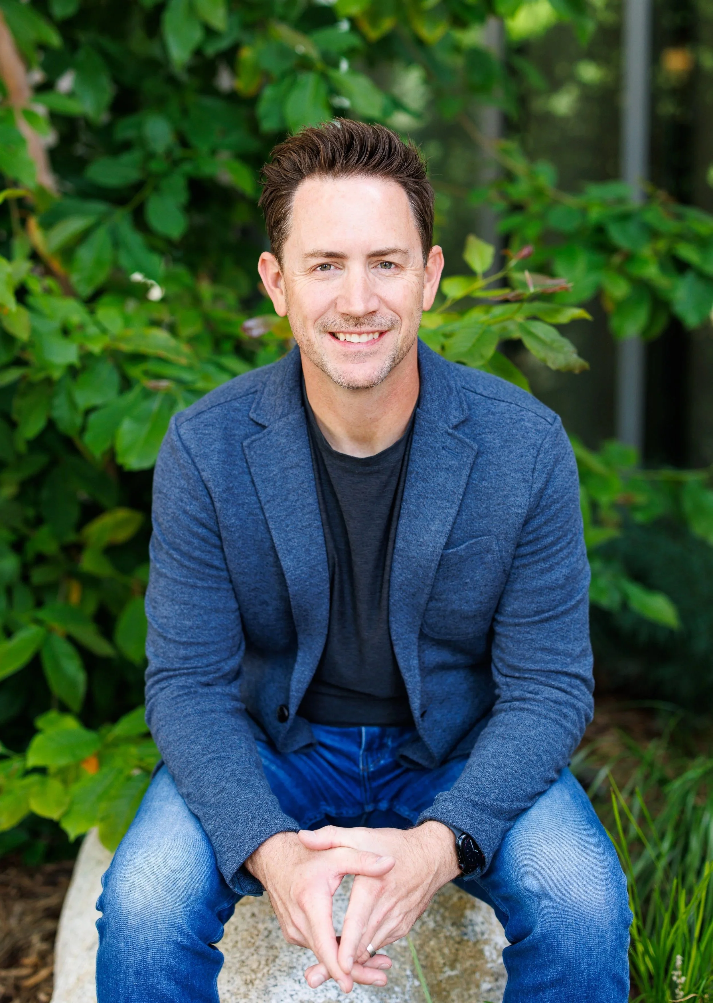 A smiling man with brown hair and blue eyes, wearing a blue blazer and jeans, sitting outdoors on a stone bench with green foliage in the background.