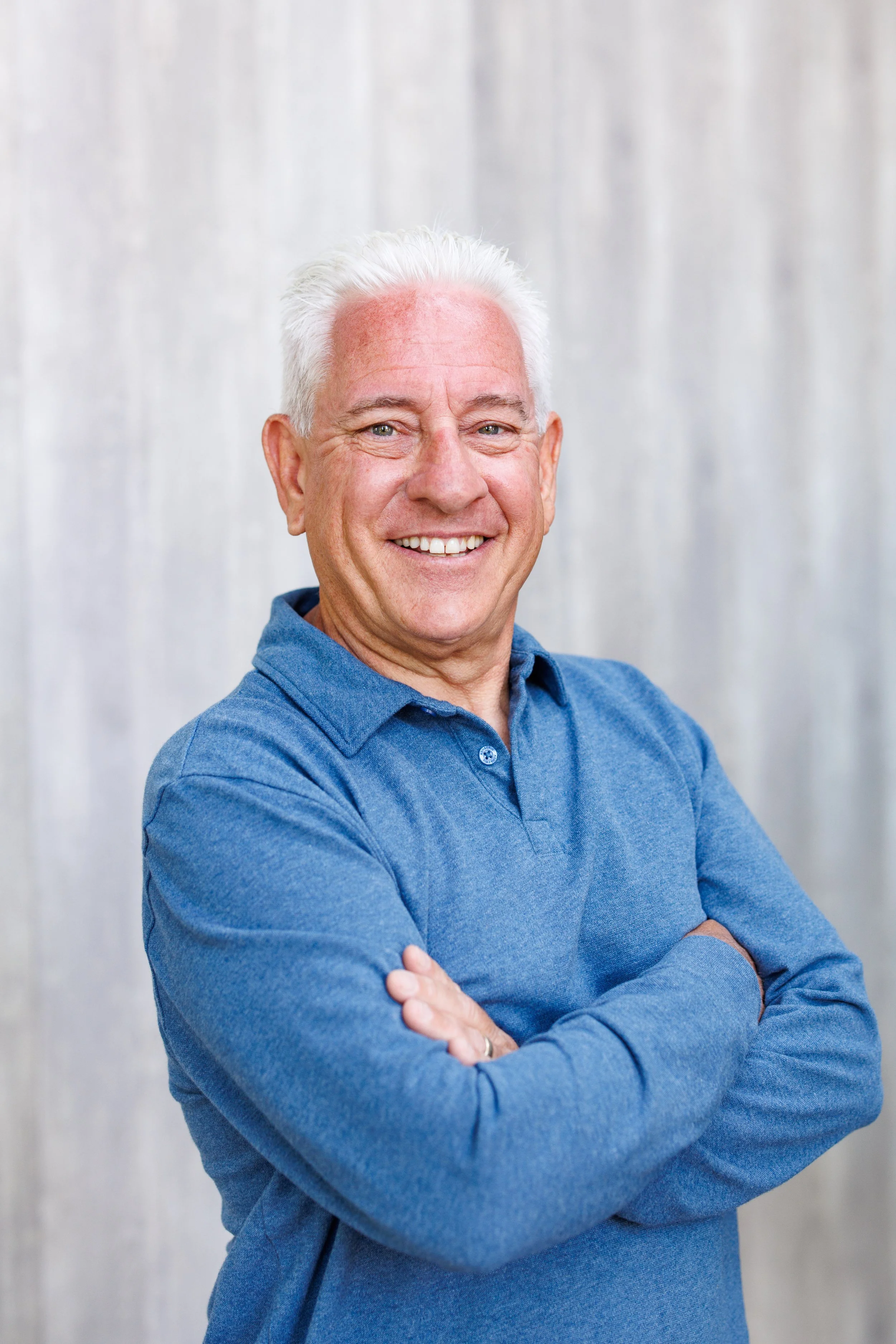 A smiling older man with white hair, wearing a blue long-sleeve shirt, standing with arms crossed in front of a light-colored background.