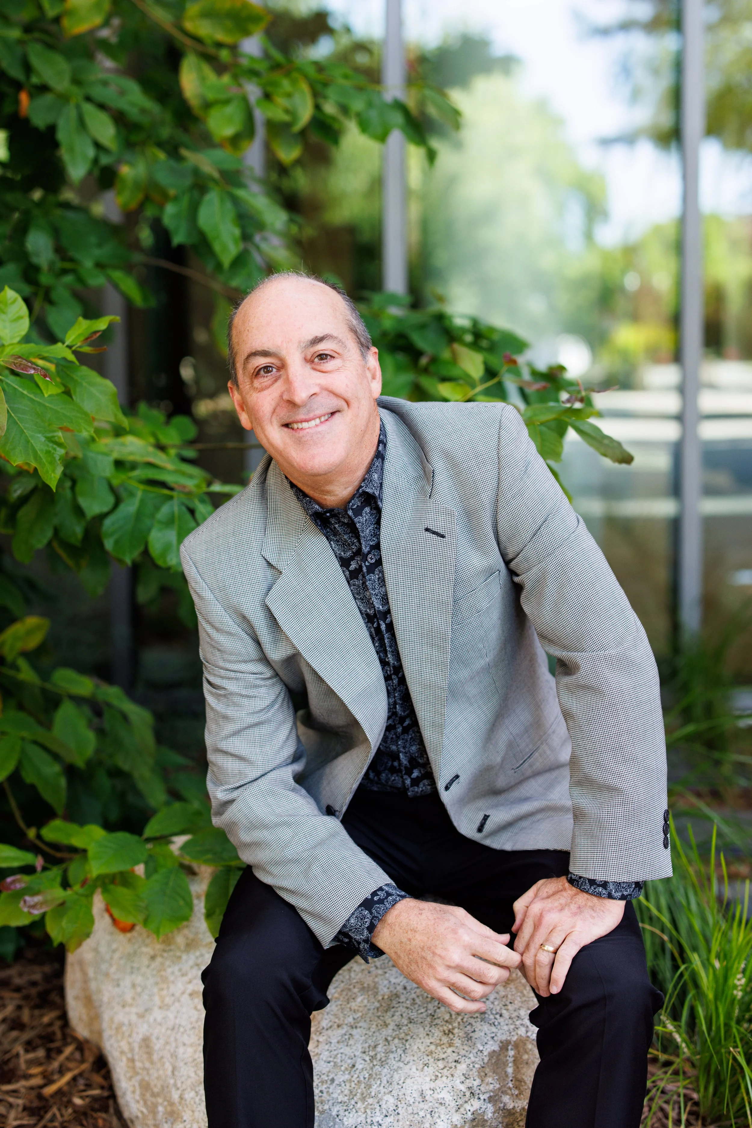 A smiling man with a receding hairline and wearing a light grey blazer over a patterned shirt, sitting outdoors on a large stone, surrounded by green plants and trees, with glass windows reflecting the outside scenery.