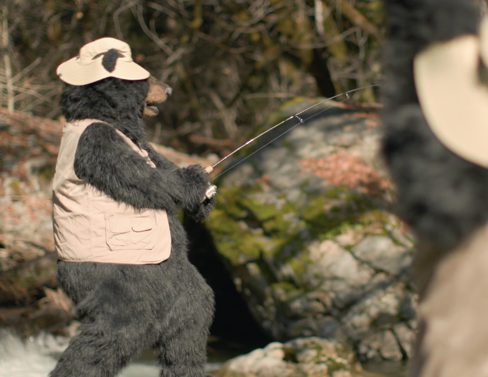 A person in a bear costume wearing a fishing hat, holding a fishing rod, fishing in a natural outdoor setting with rocks and trees.
