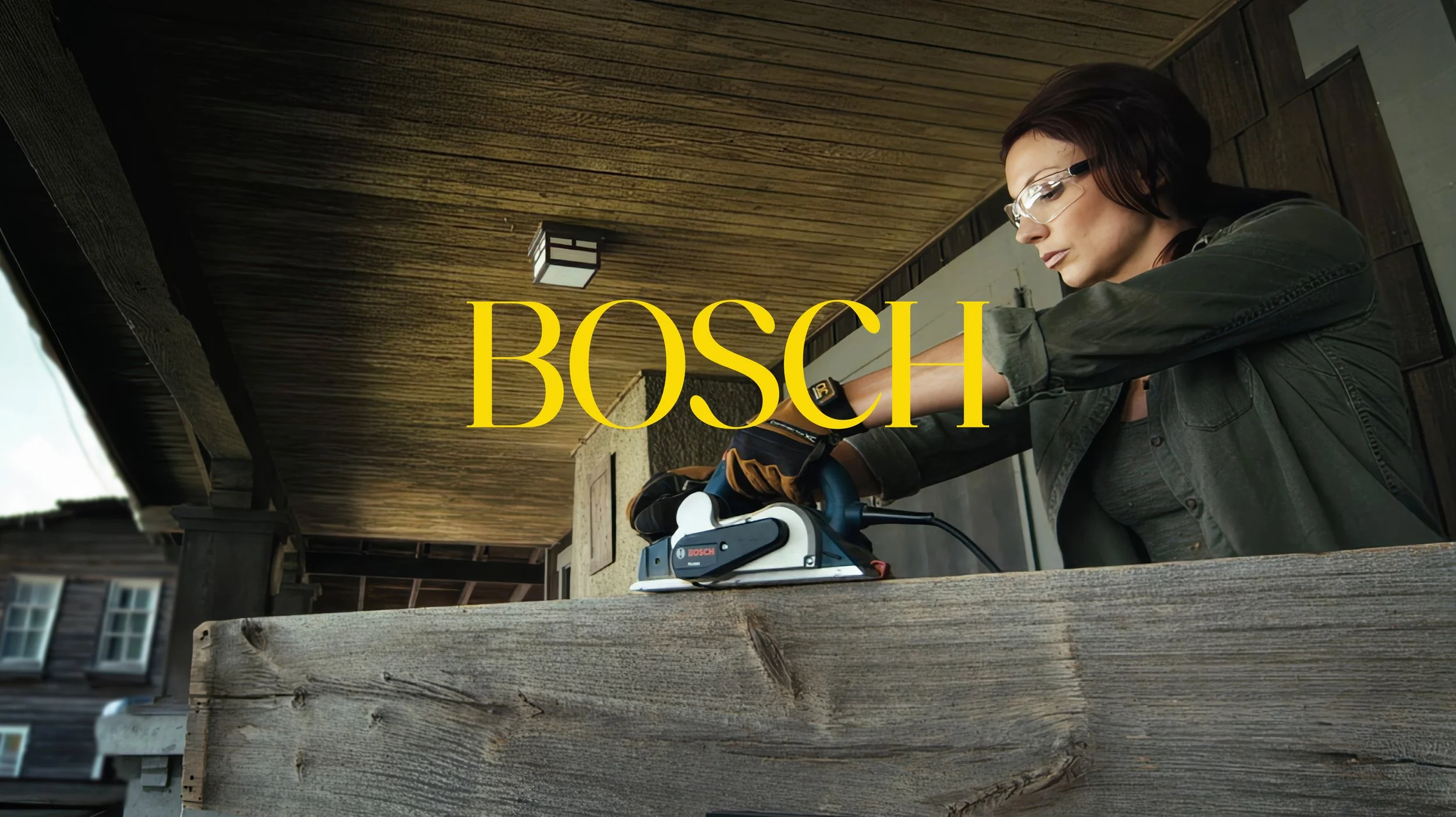A woman with glasses and a green jacket operating a Bosch cordless power planer on a wooden surface, outside of a house with a porch.