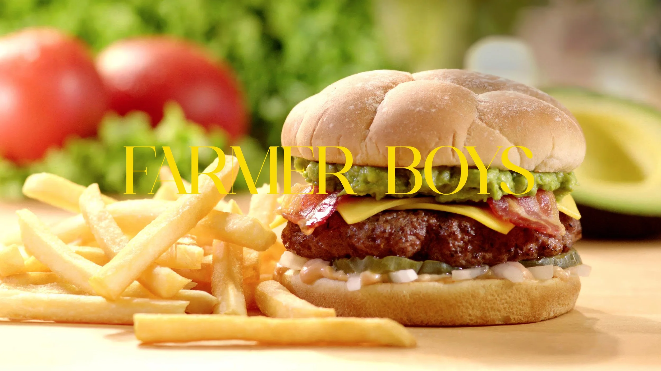 A hamburger with bacon, cheese, guacamole, and pickles on a bun, served with French fries, with tomatoes and lettuce in the background, and the text 'FARMER BOYS' over the burger.