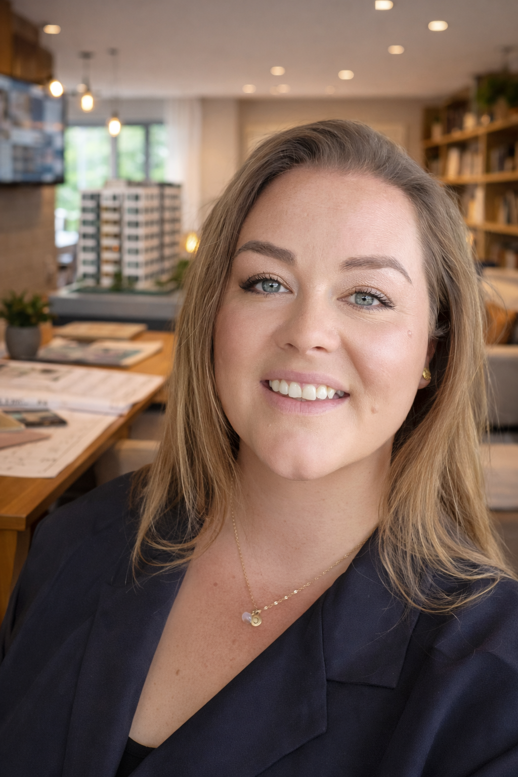 A woman with blonde hair and blue eyes smiling at the camera in a modern, well-lit room with bookshelves and a model of a building in the background.