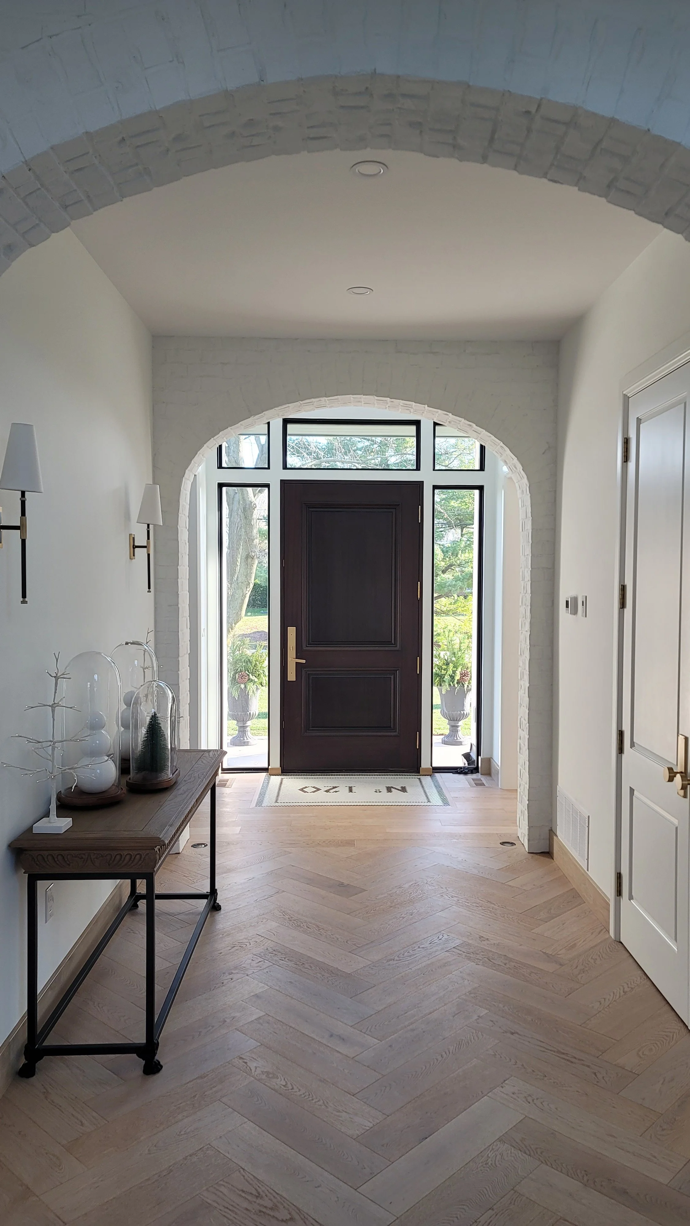 Entryway with dark front door, white brick archways, light wood herringbone flooring, and wall-mounted lamps, with potted plants visible through glass sidelights.