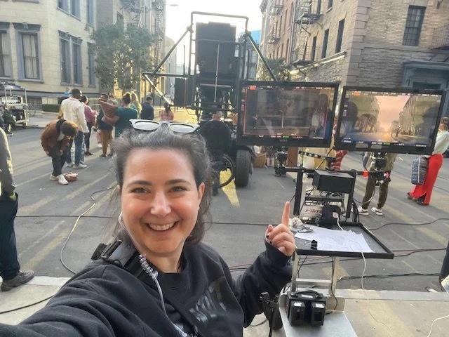 A woman on a film set taking a selfie with camera equipment and monitors behind her, people working in the background with city buildings.