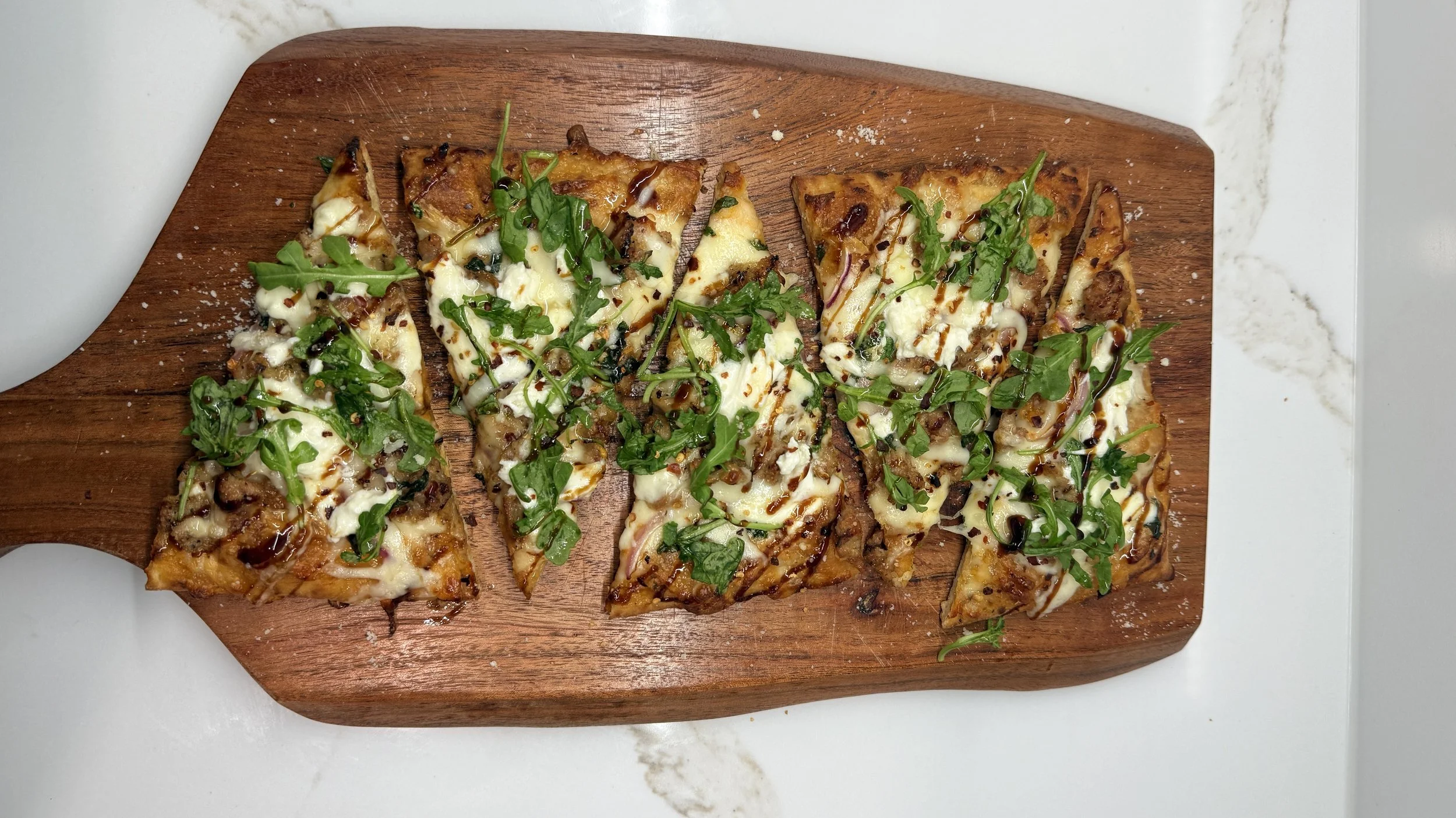 Slices of flatbread pizza topped with cheese, arugula, and drizzled with balsamic glaze on a wooden serving board.