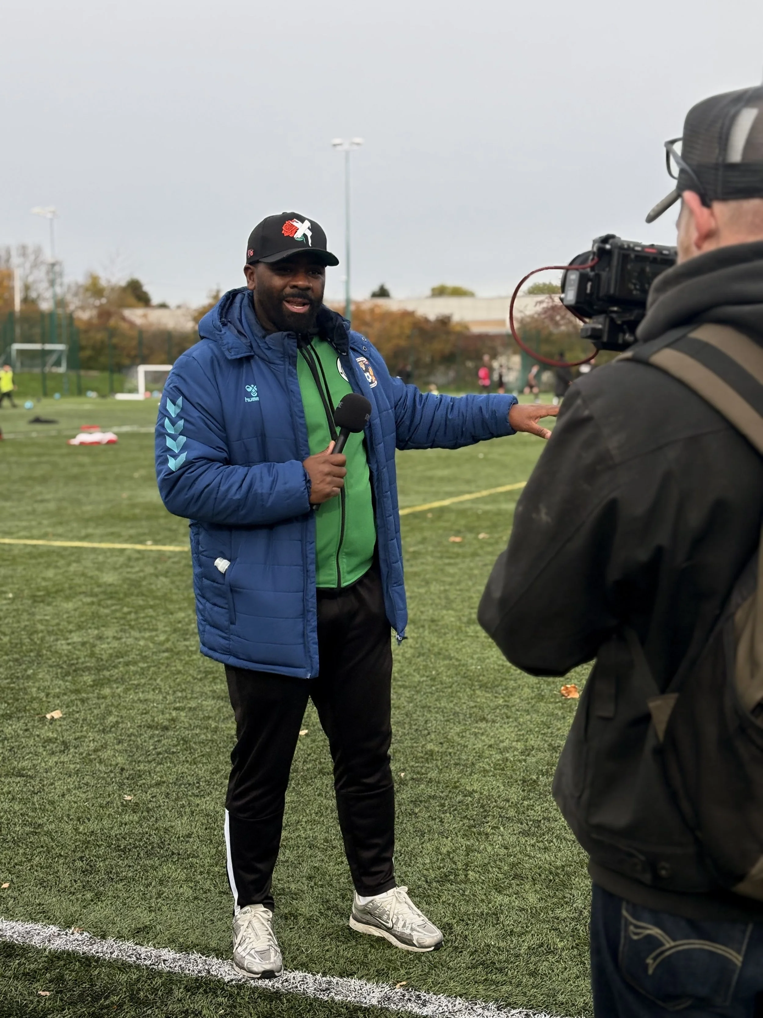 A man in a blue jacket and black pants holding a microphone is being recorded by a person with a camera on a football field. Piles of soccer balls are visible in the background.